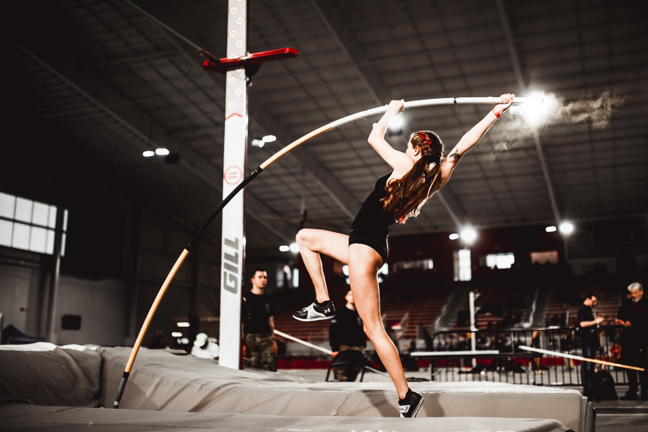 A female athlete in black athleticwear competing in pole vault in an indoor arena, with people observing in the background.