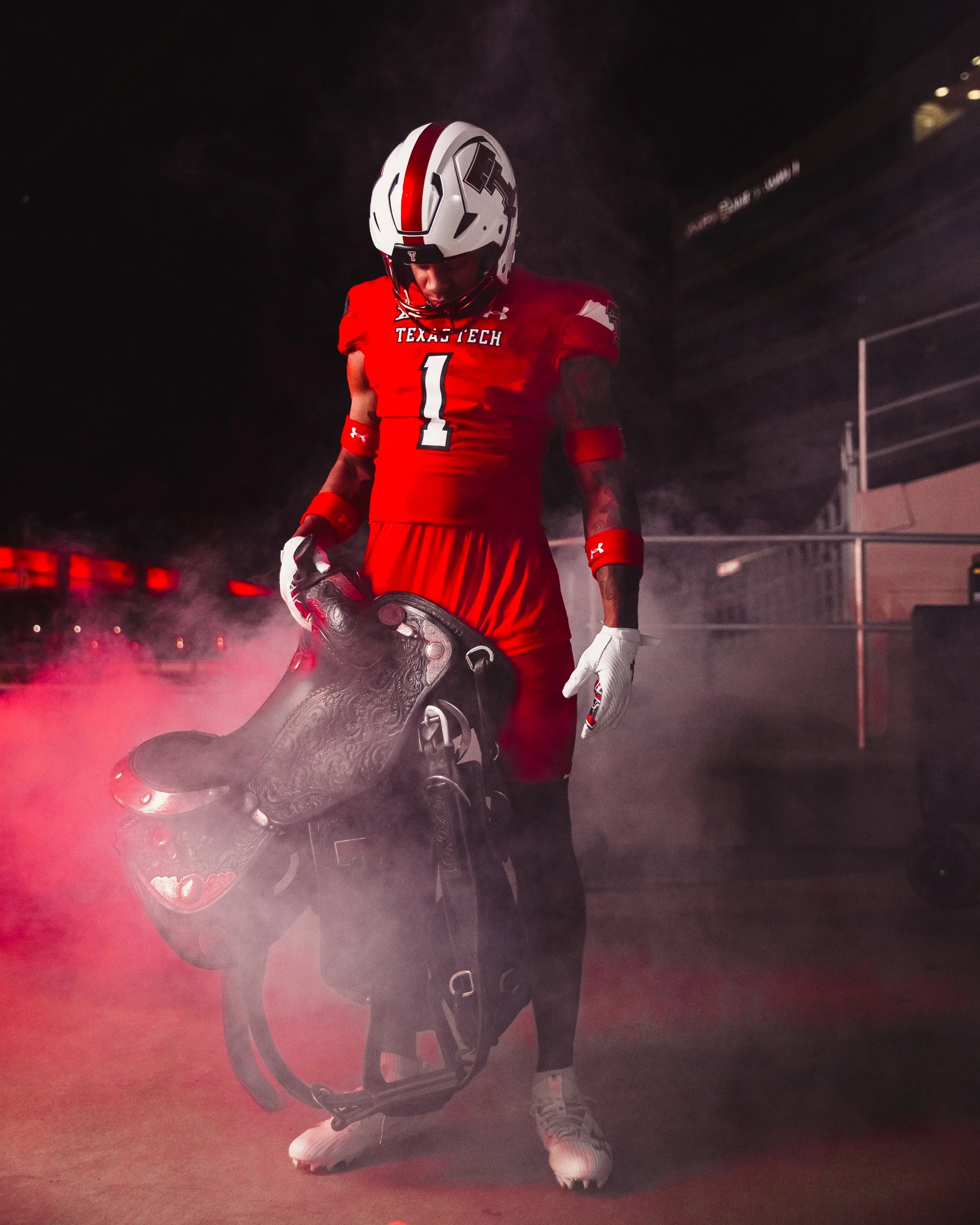 A football player from Texas Tech University wearing a red uniform and white helmet stands with head bowed, holding a helmet in one hand, surrounded by red smoke.