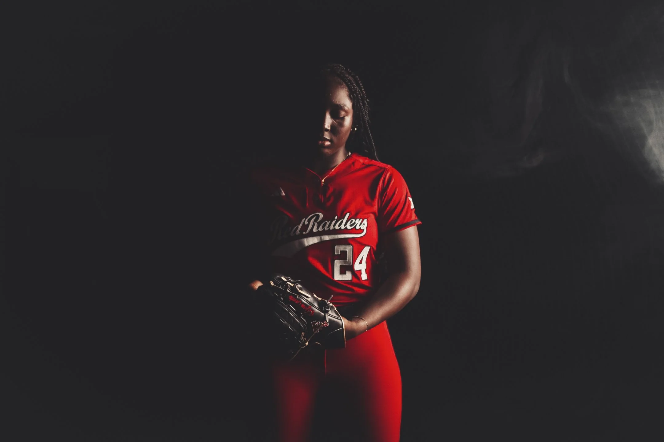 A female softball player in a red jersey with the number 24 and 'Road Raiders' written on it, standing with a glove on her left hand in a dark setting with dramatic lighting.