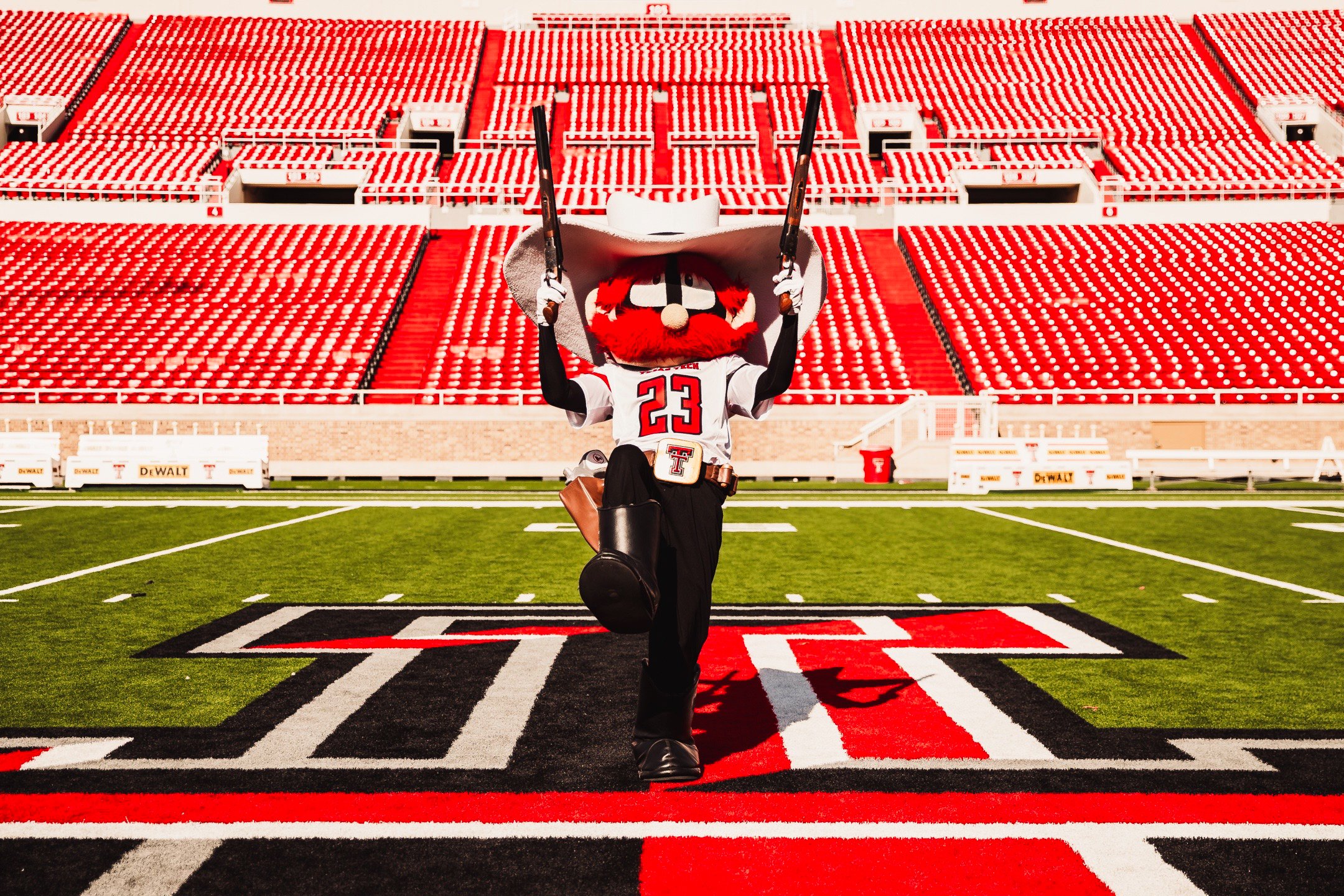 University of Texas at Austin mascot, Hook 'Em, dressed in football uniform with jersey number 23, holding two swords, walking on the football field with empty stadium seats in the background.