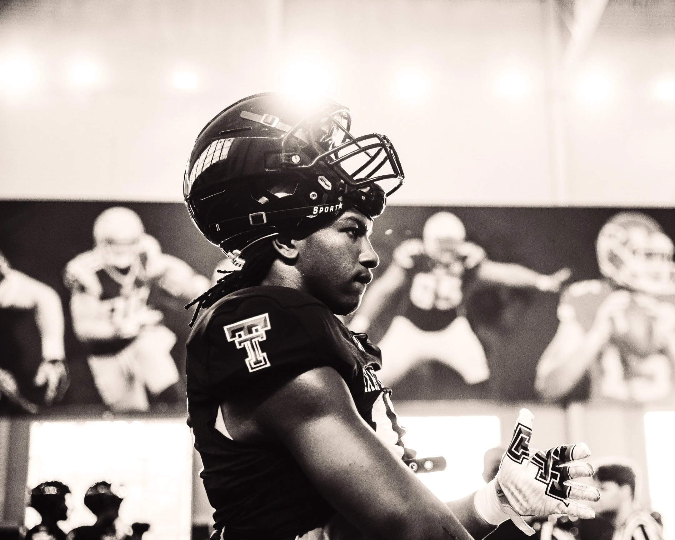 Black and white photo of a football player in uniform and helmet inside a locker room, with a wall mural of football players in the background.