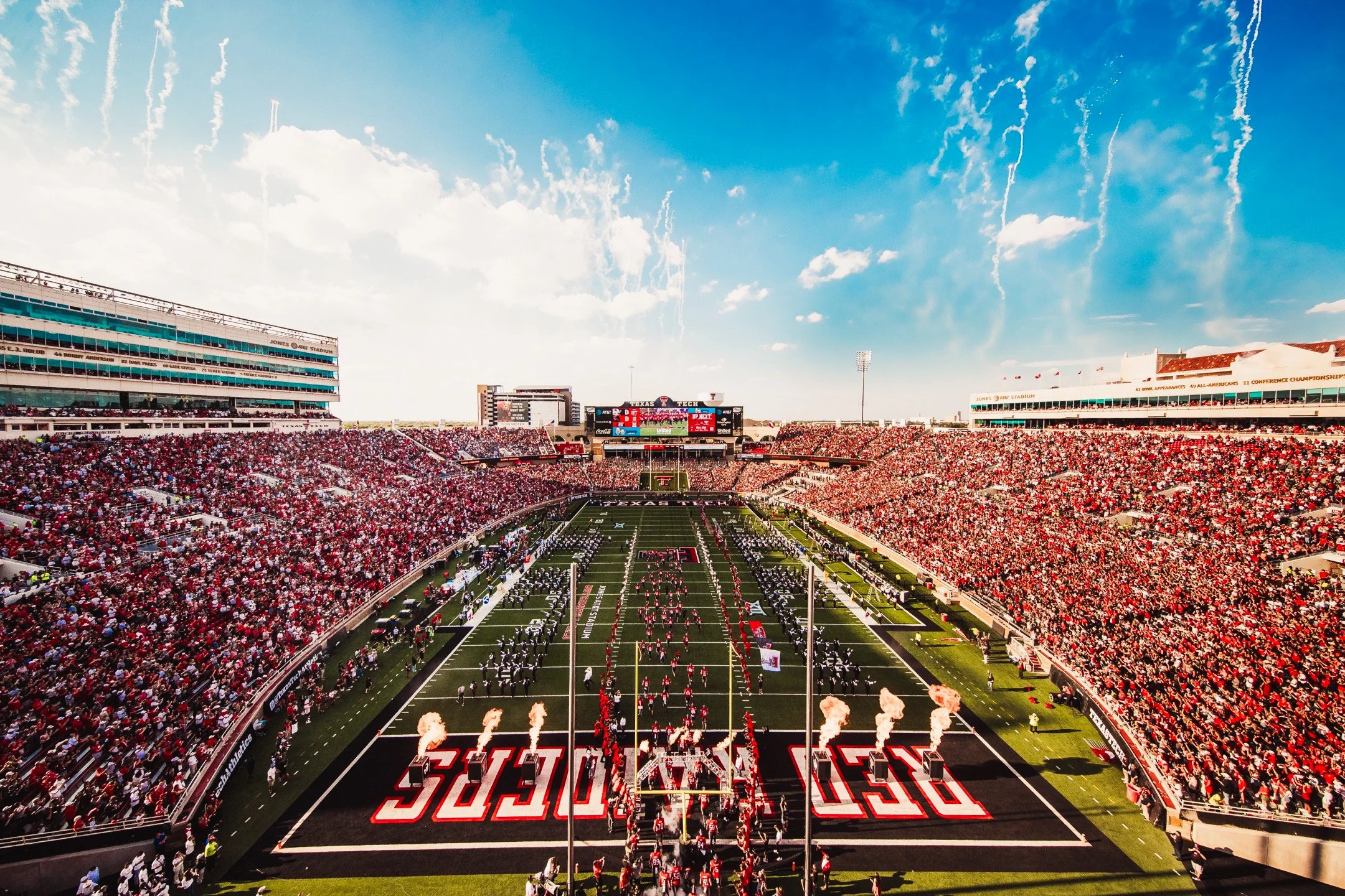 A packed football stadium during a game, with players on the field and fans in the stands, under a bright blue sky.
