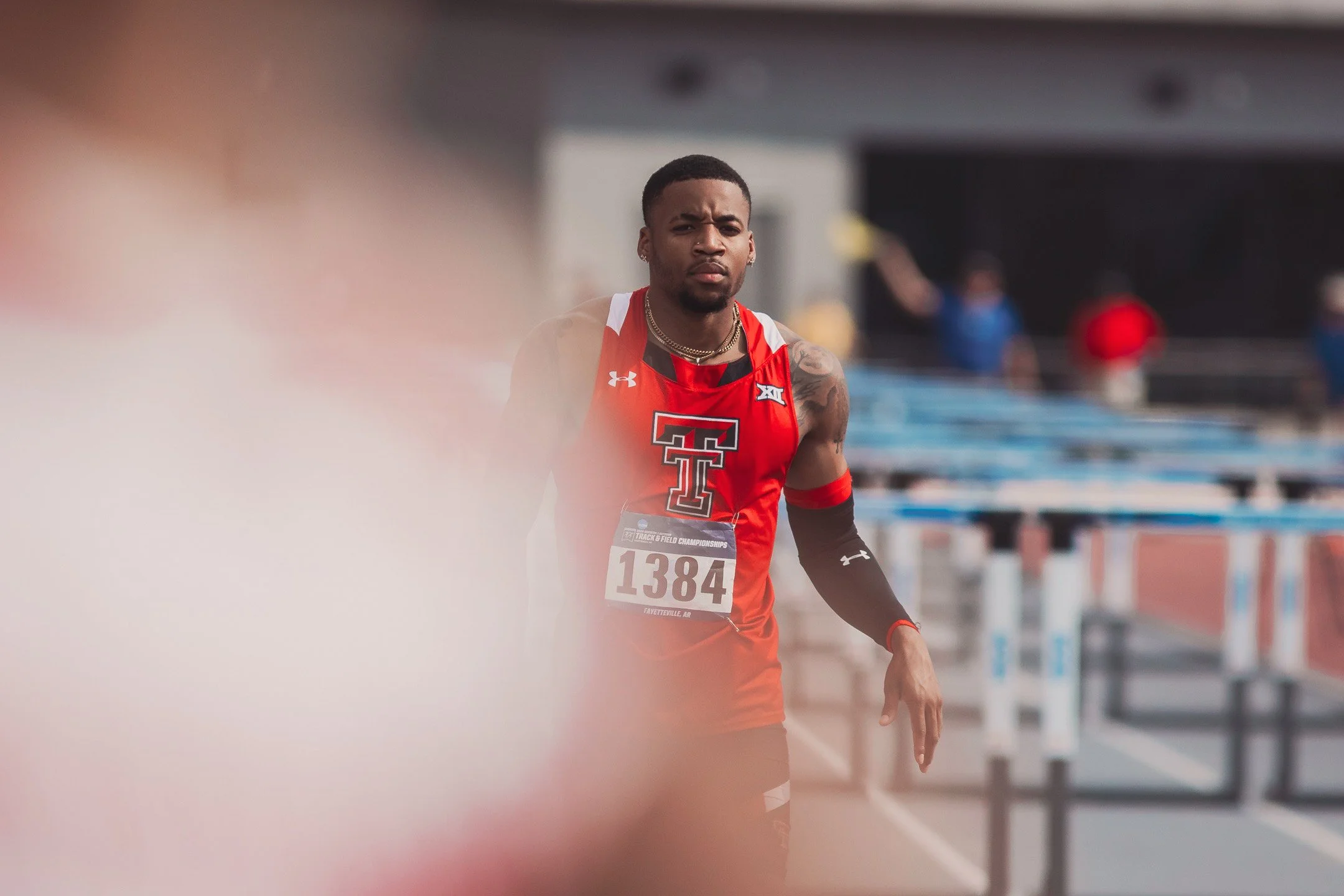 A male athlete wearing a red sleeveless sports shirt with the Texas Tech logo at a track and field event. He has a race bib number 1384, black arm sleeves, and is standing on a running track.
