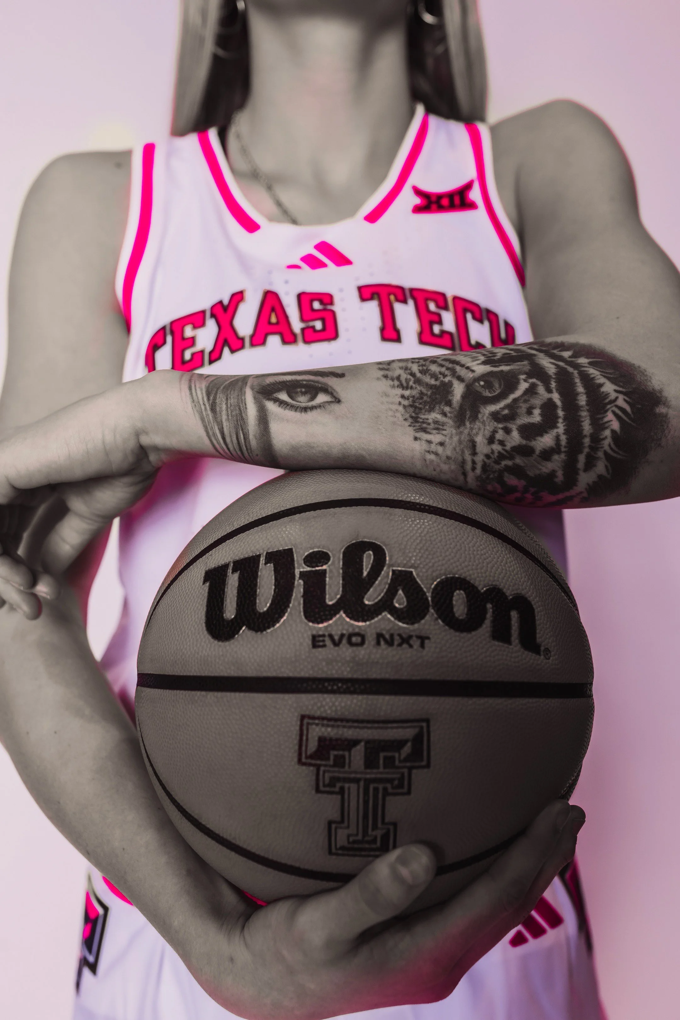 A female basketball player wearing a white Texas Tech jersey with pink accents, holding a Wilson basketball in front of her. She has a tattoo of a tiger and a human face on her arm.