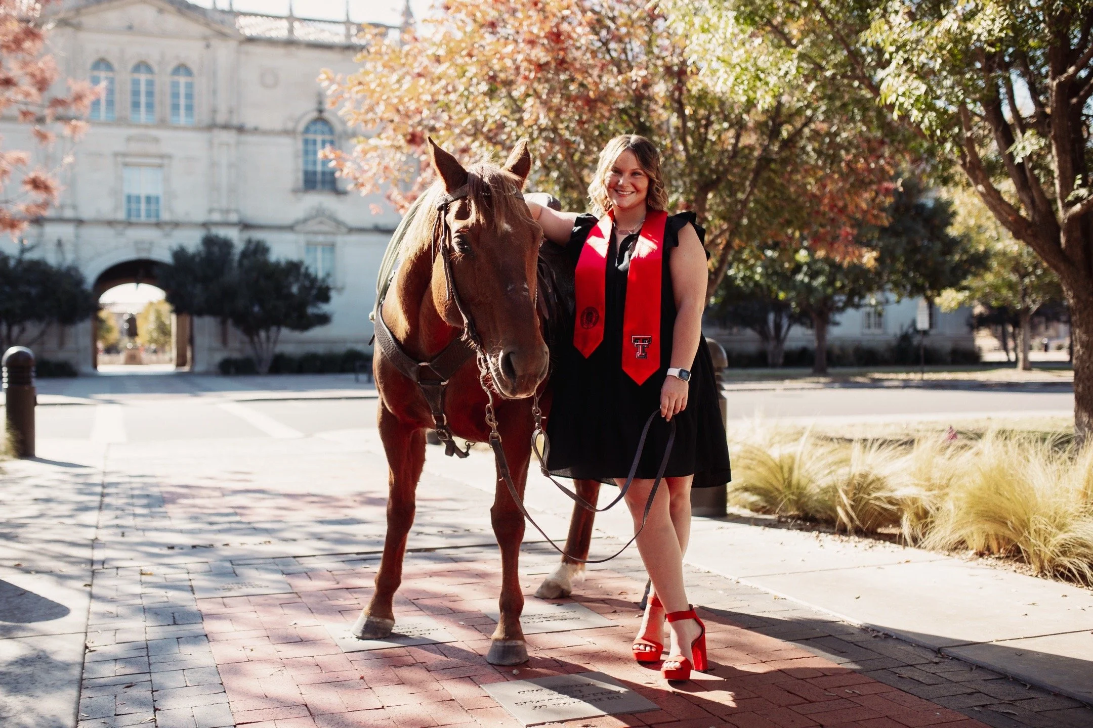 A woman in a black dress with a red graduation stole and red high heels standing next to a brown horse on a city sidewalk with trees and a historic building in the background.