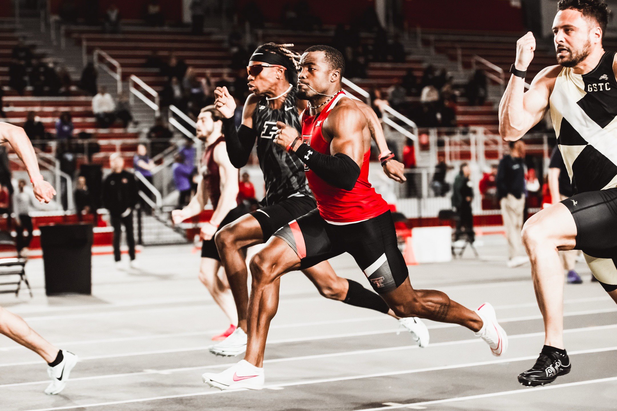 Track athletes sprinting at a competition in an indoor arena with spectators in the background.