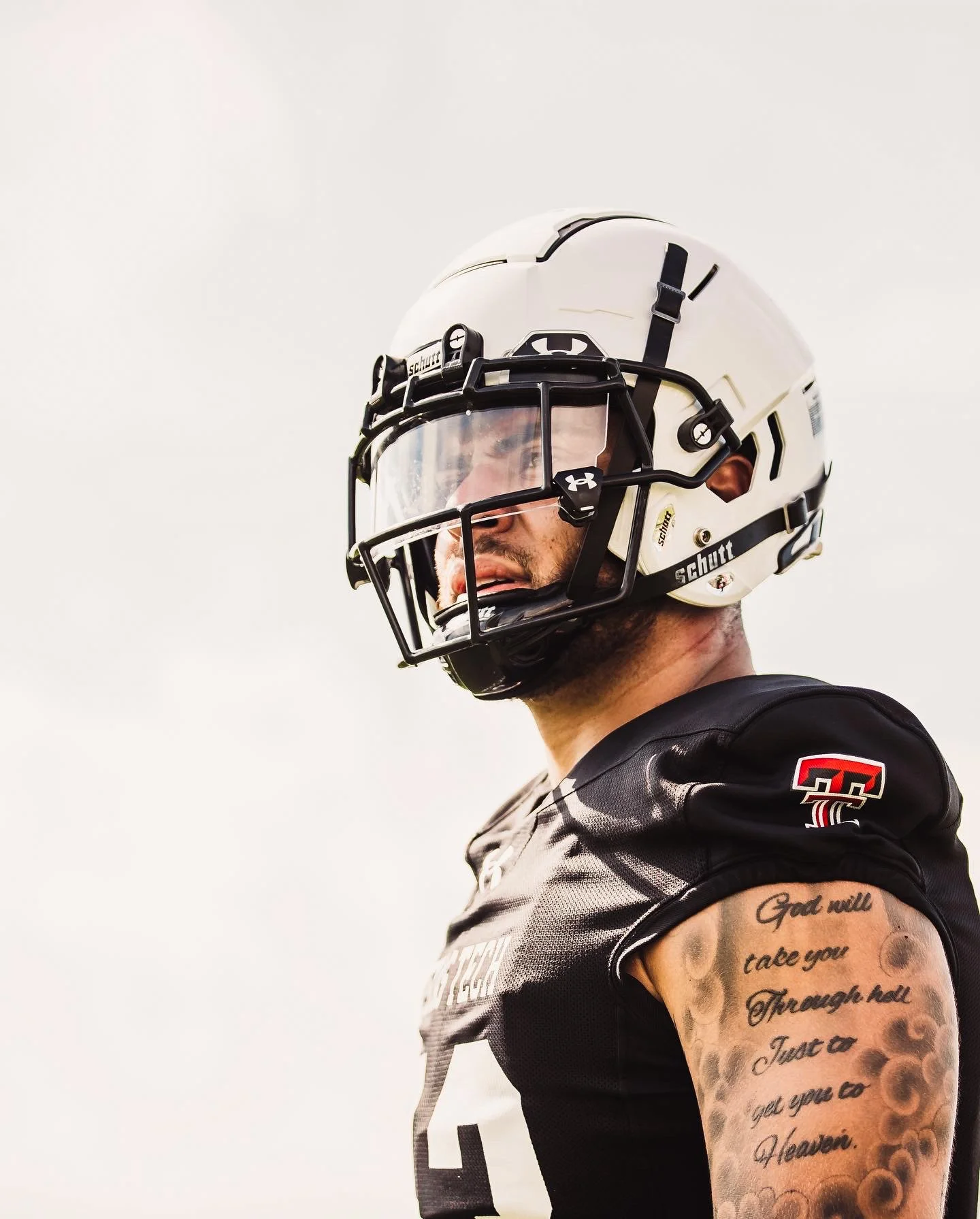 A football player wearing a white helmet with a faceguard, black jersey with red and white accents, and tattooed arm, standing outdoors.
