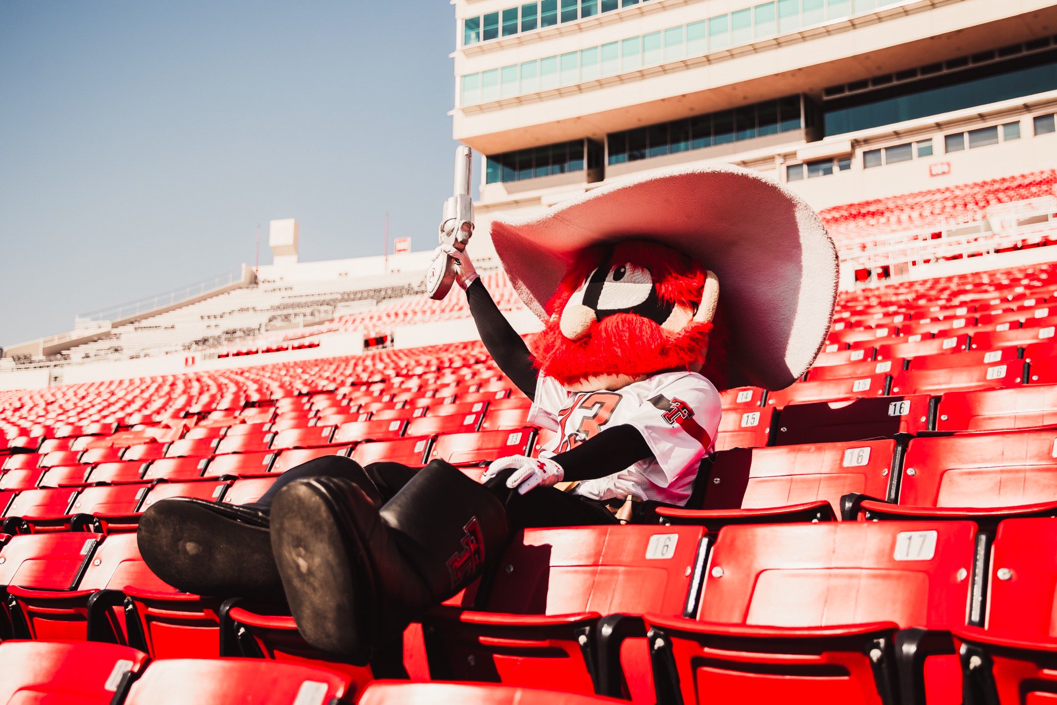 A football mascot with a large red beard, big black eyes, a big wide-brimmed hat, and a jersey, sitting alone in red stadium seats, holding a sword in one hand, with a modern stadium background under a clear sky.
