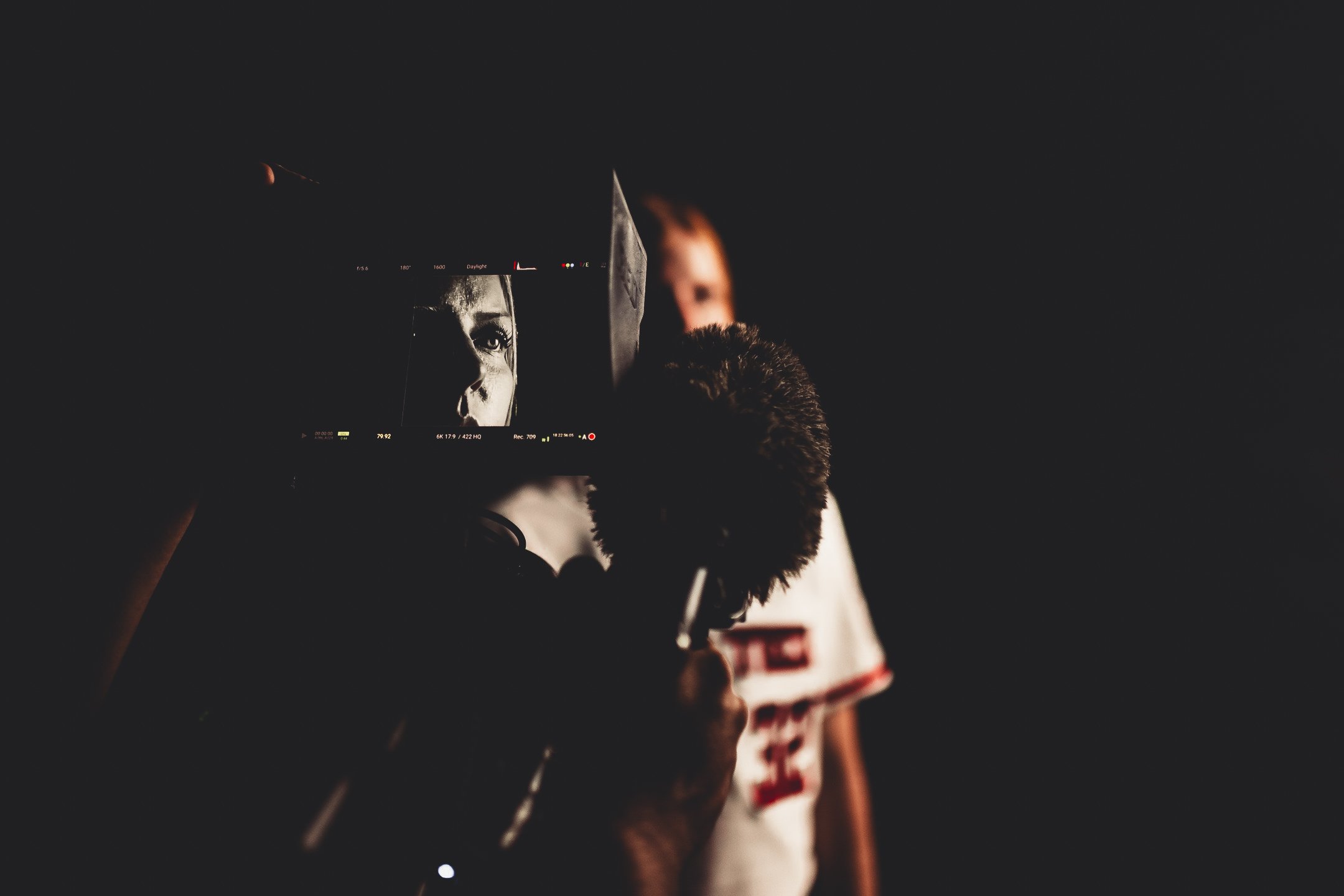 A person filming a movie scene with a camera, capturing a woman in a sports jersey standing against a dark background.