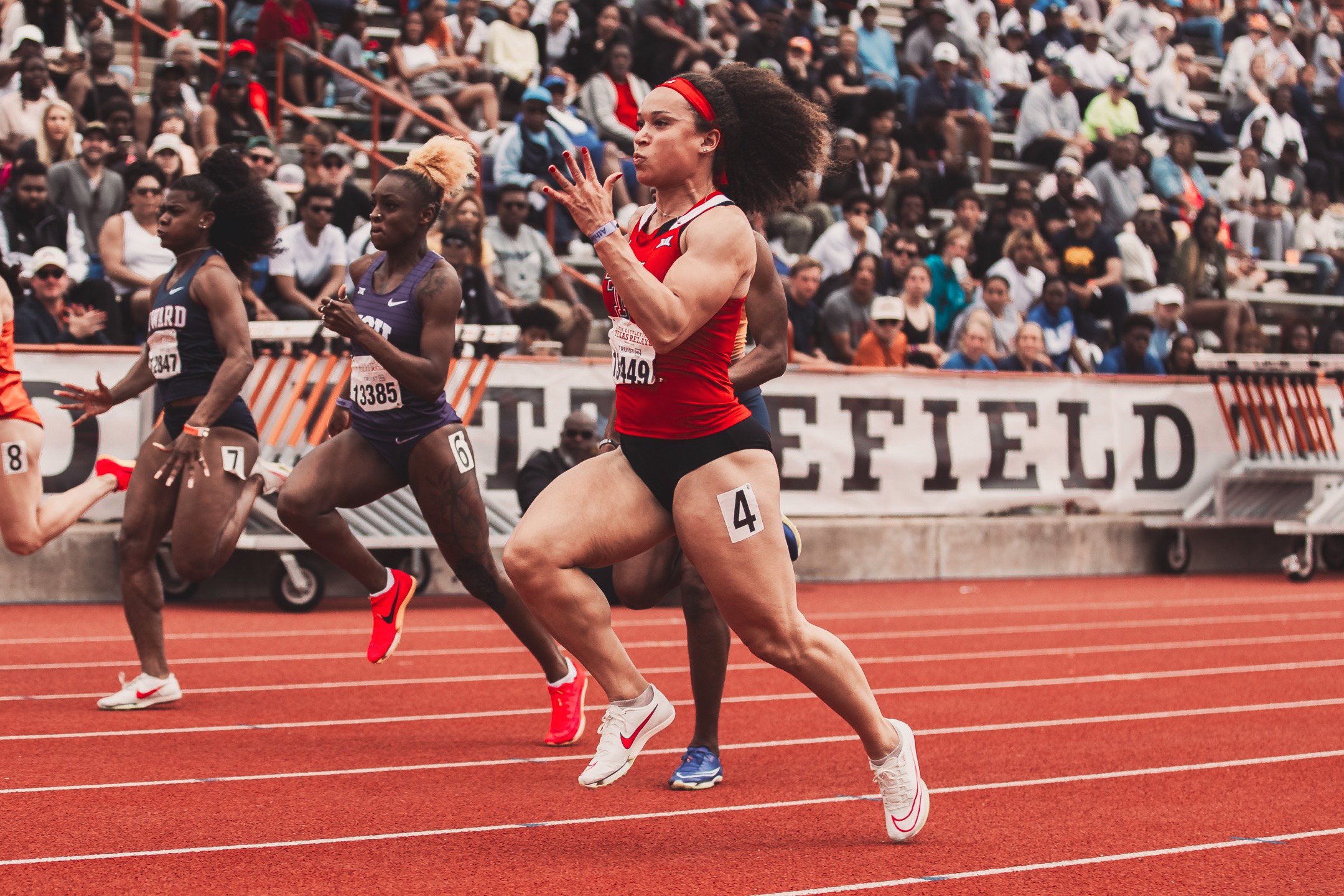 Female athletes running on a track during a race, with a large crowd of spectators in the background.