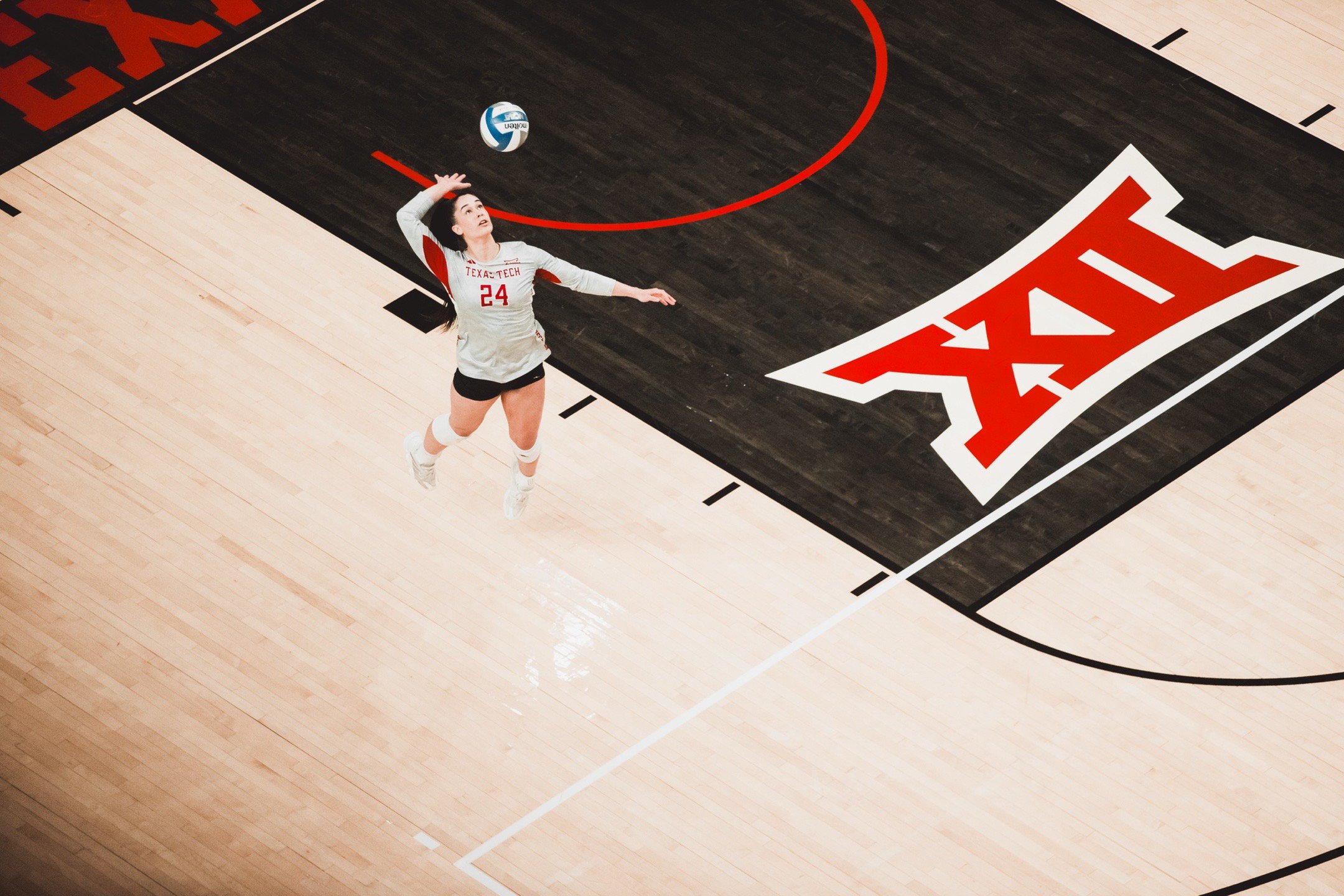 A female volleyball player in a white jersey with the number 24 and 'TEXAS TECH' on it, jumping to hit a volleyball on a basketball court with the Big 12 logo.