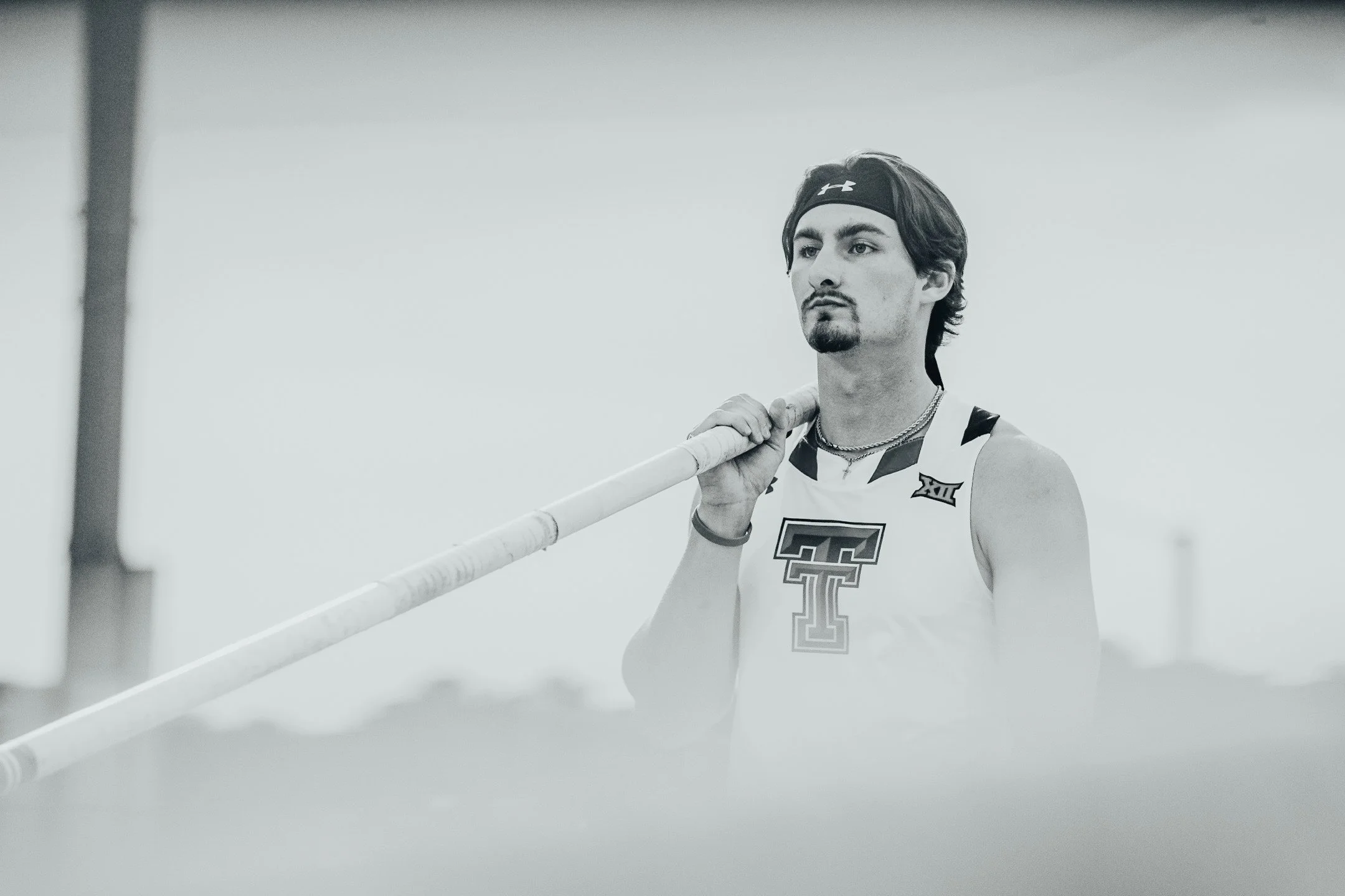 A male track athlete holding a pole over his shoulder, wearing a sleeveless sports jersey with a large double T on the front, a headband, and necklaces.