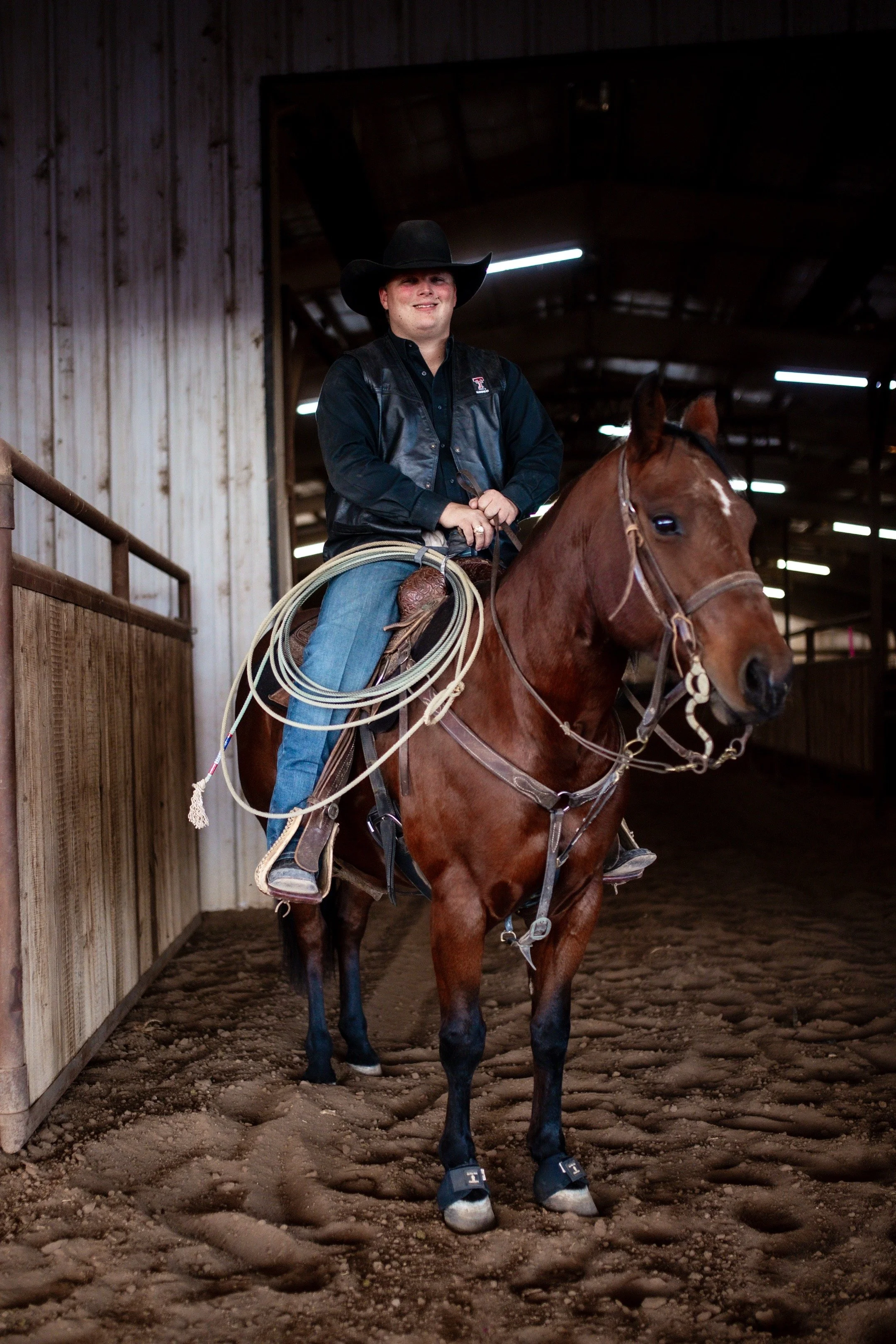 A person wearing a black cowboy hat, black vest, and jeans riding a brown horse inside a barn.