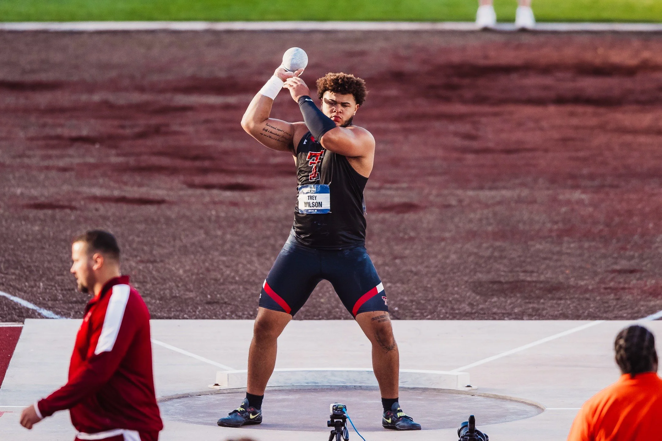Young male athlete preparing to throw a shot put during a track and field event, standing on a concrete circle with a reddish-brown track in the background, wearing a black athletic outfit and a white wrist wrap.