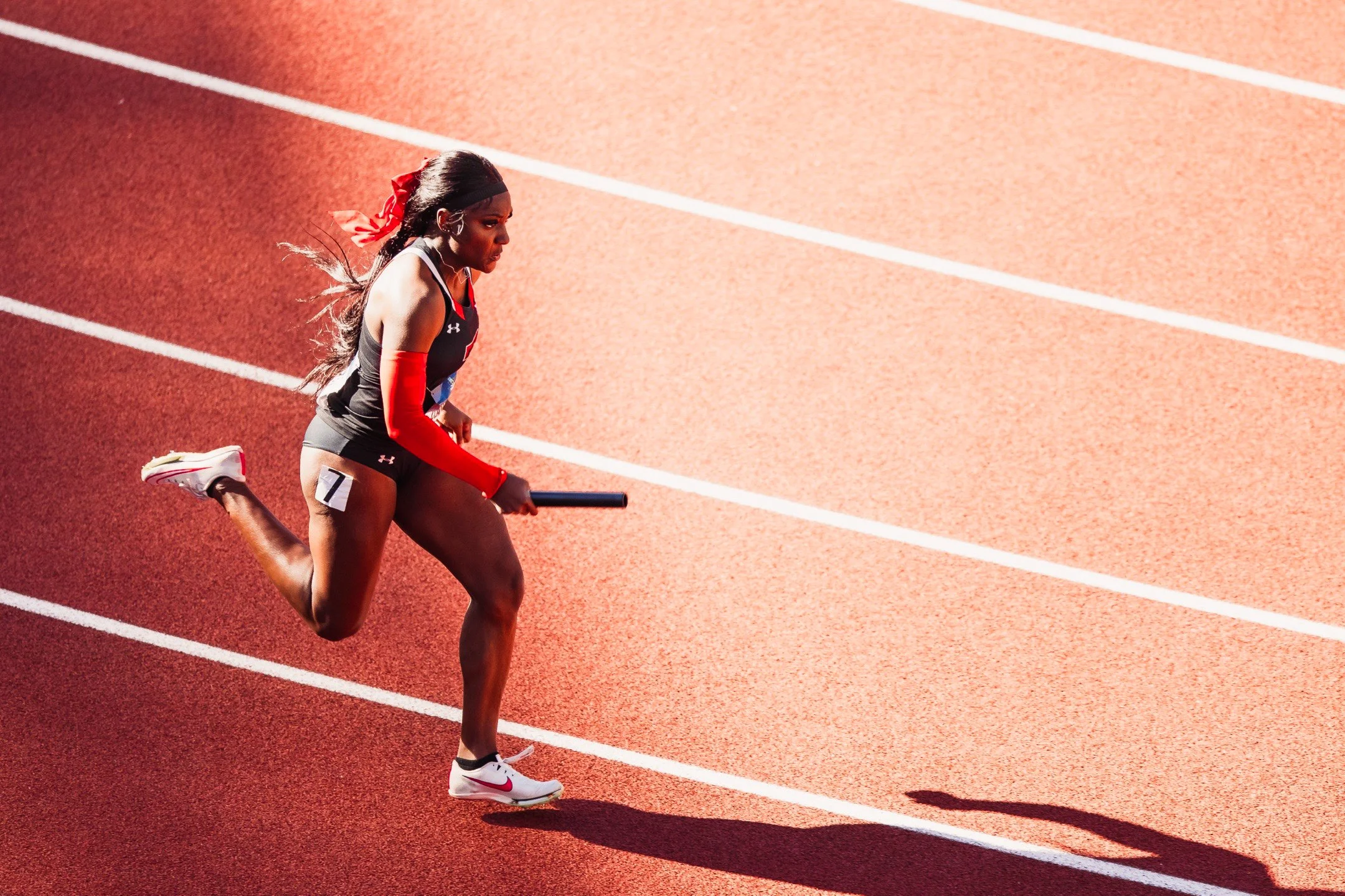 A female athlete with long hair in a ponytail, wearing a black tank top, black shorts, red arm sleeves, and white running shoes, running on a red track with white lane lines and holding a baton.
