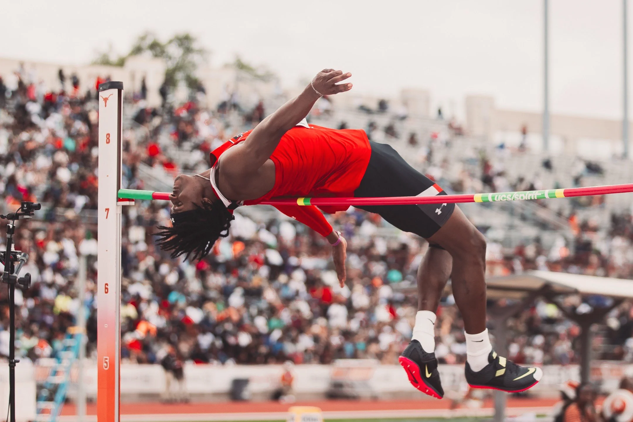 Athlete clearing a high jump bar at a track event, over a background of a crowd in the stadium.