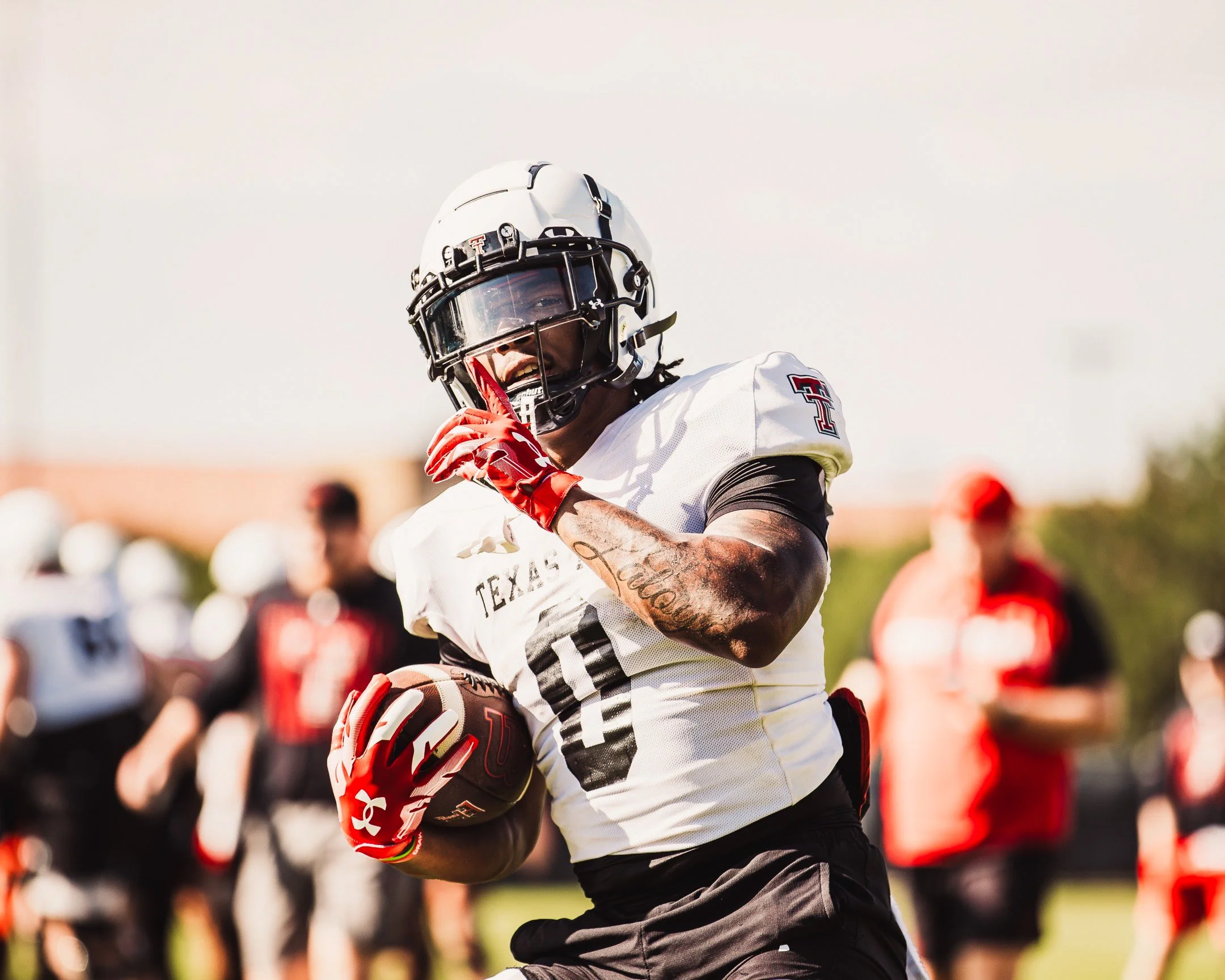 An American football player wearing a white Texas Tech jersey, black pants, red gloves, and a helmet holding a football while running on the field during a game.