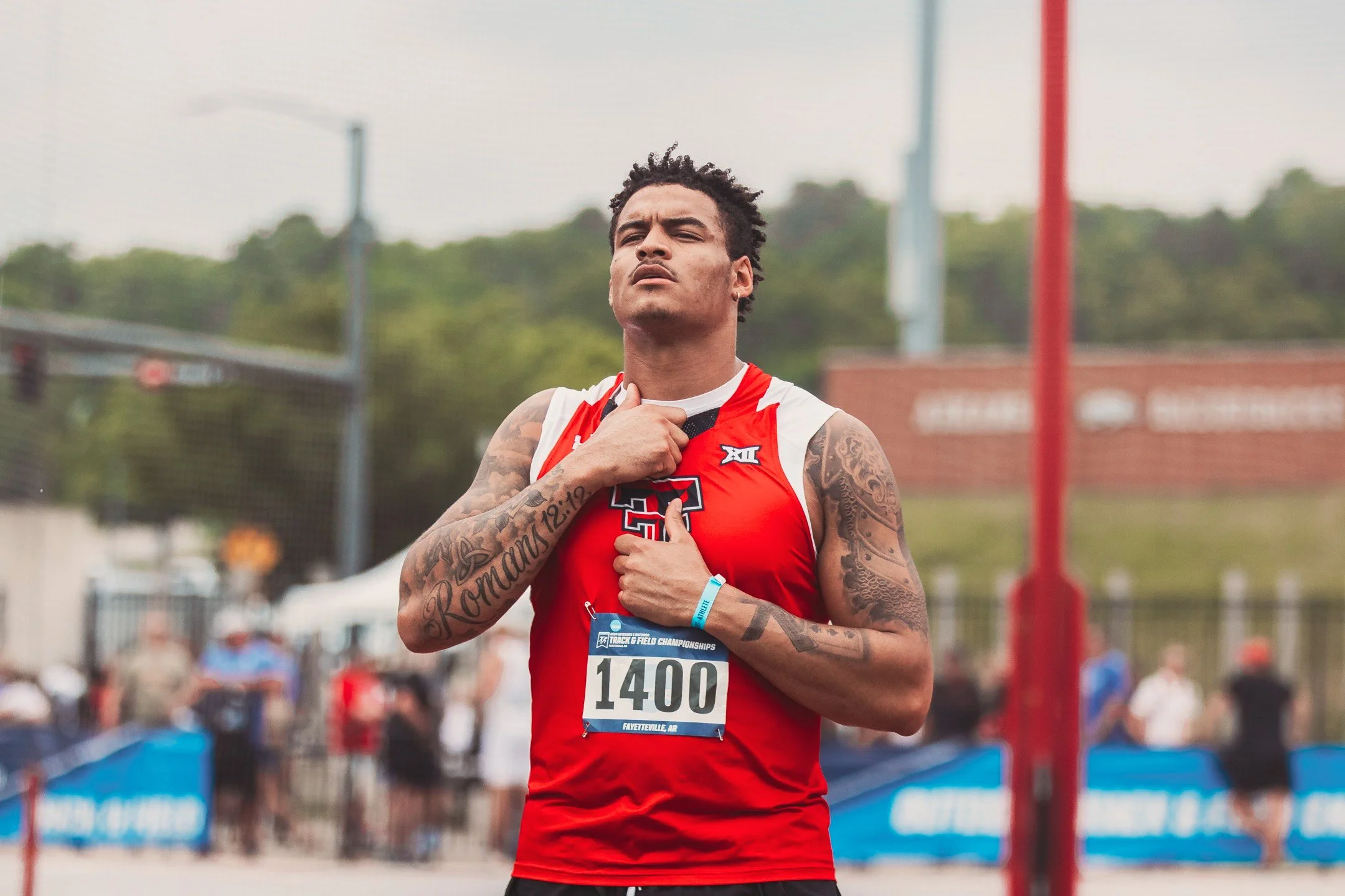 Male athlete in red sleeveless jersey with bib number 1400, holding his hand over his chest, standing on a track or field during a sports event.