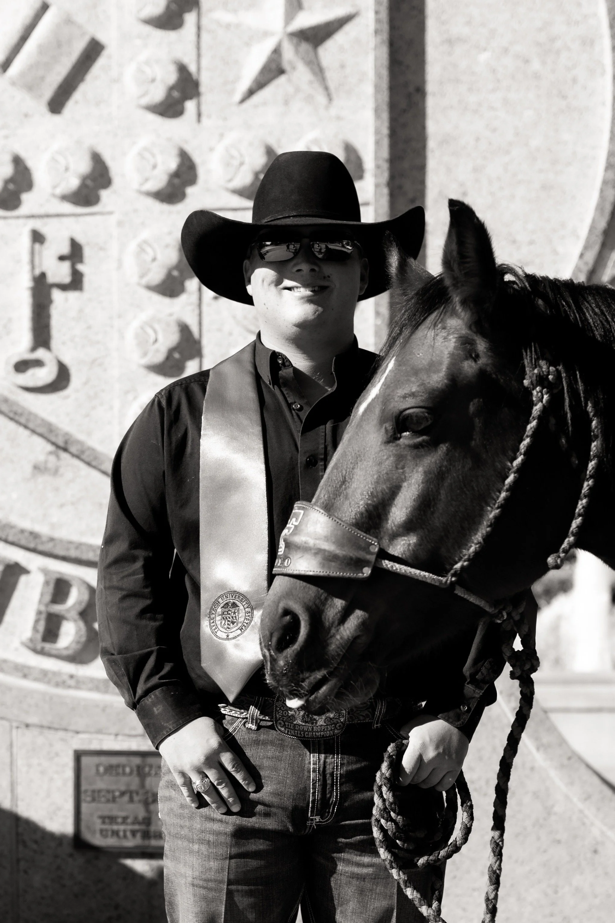A man wearing a cowboy hat, sunglasses, and a sash stands next to a horse in front of a large wall with a star emblem, in black and white.