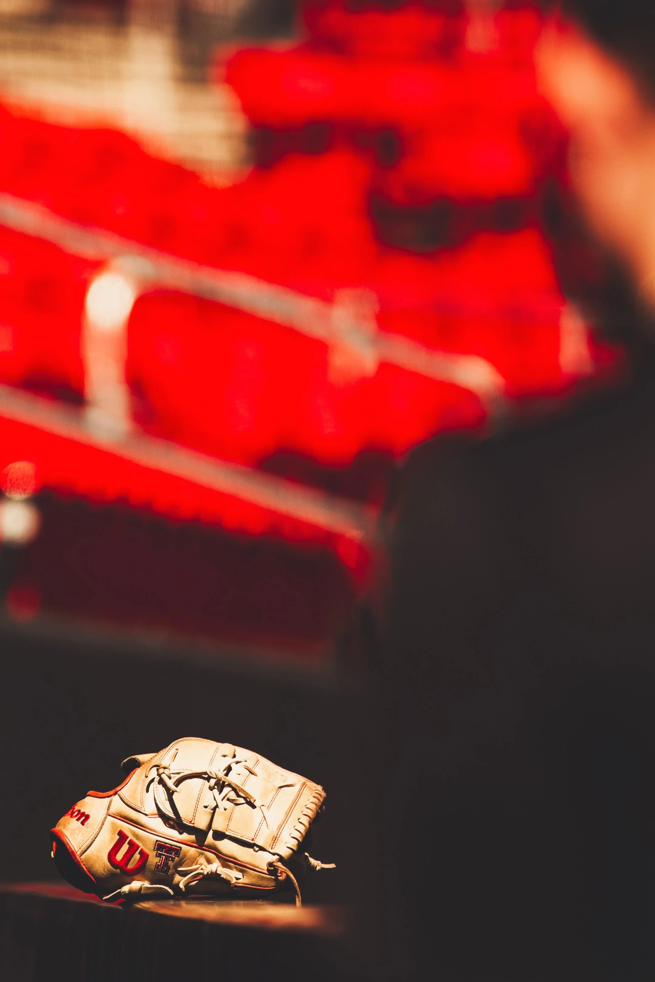 A baseball glove with the University of Texas logo on it, placed on a wooden surface with a blurred red stadium seat background.
