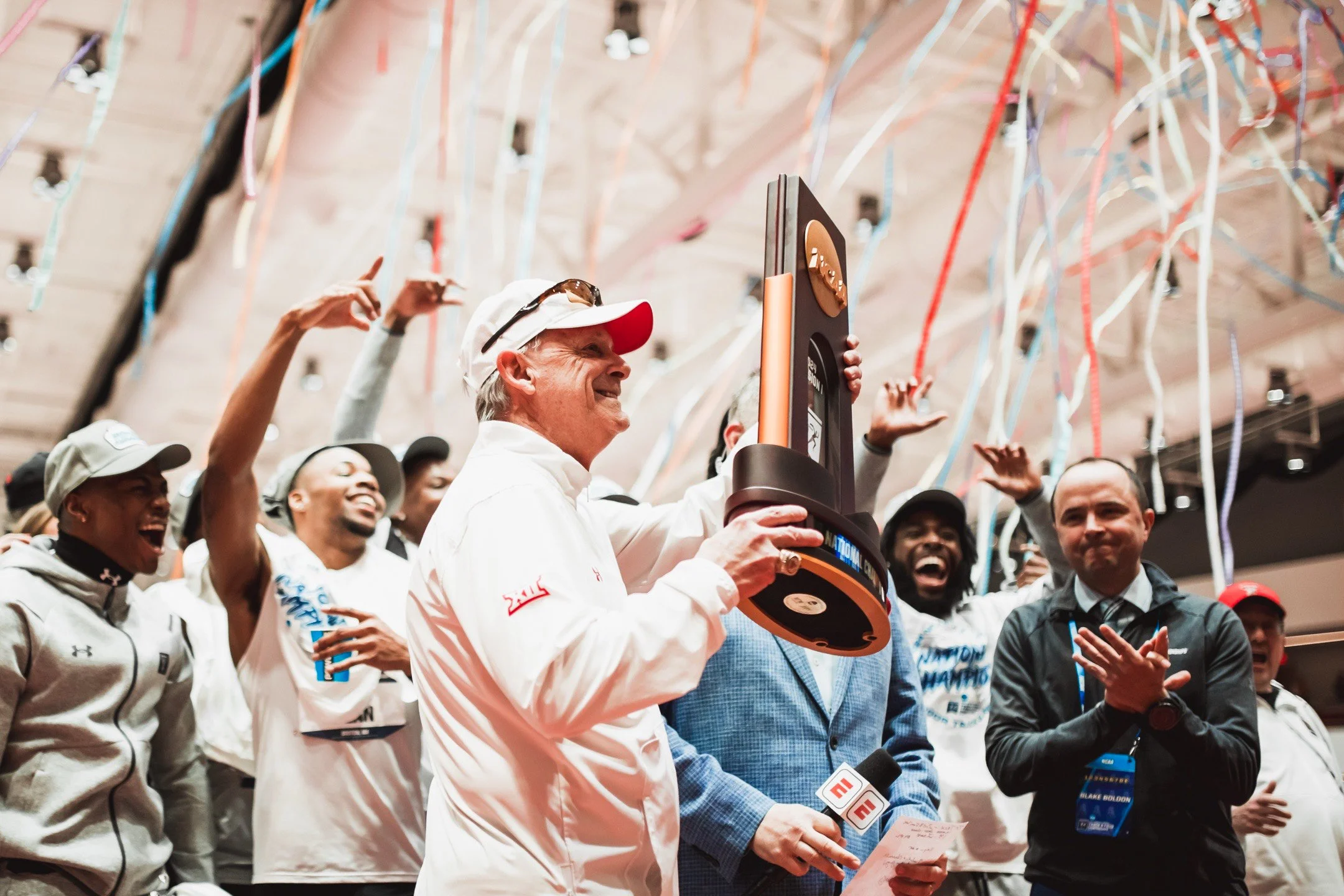 A man in a white jacket and white cap holding a large trophy while other people celebrate around him, some clapping and smiling, with colorful streamers hanging from the ceiling.