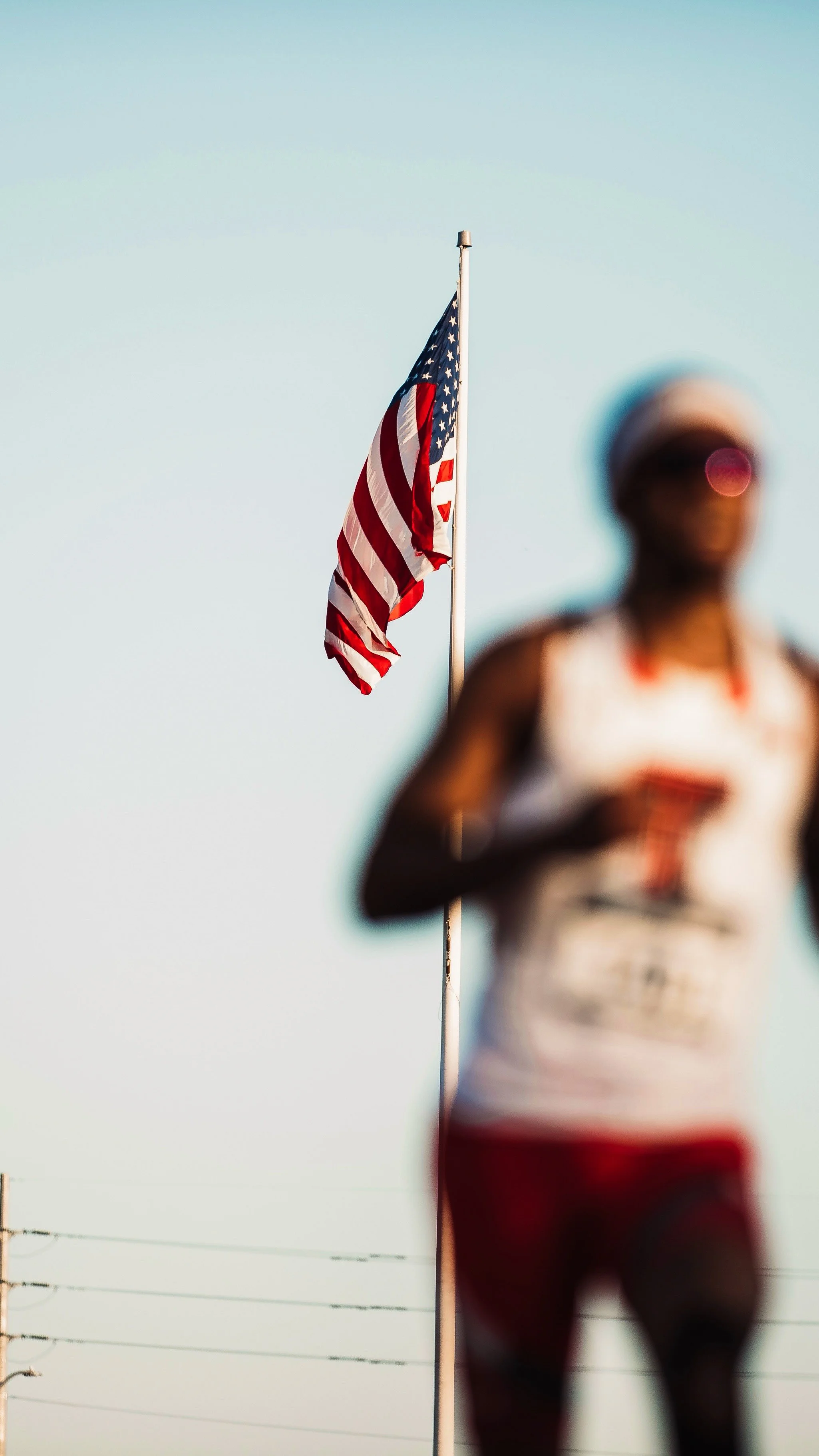 A blurry runner in athletic gear with an American flag flying on a pole in the background.