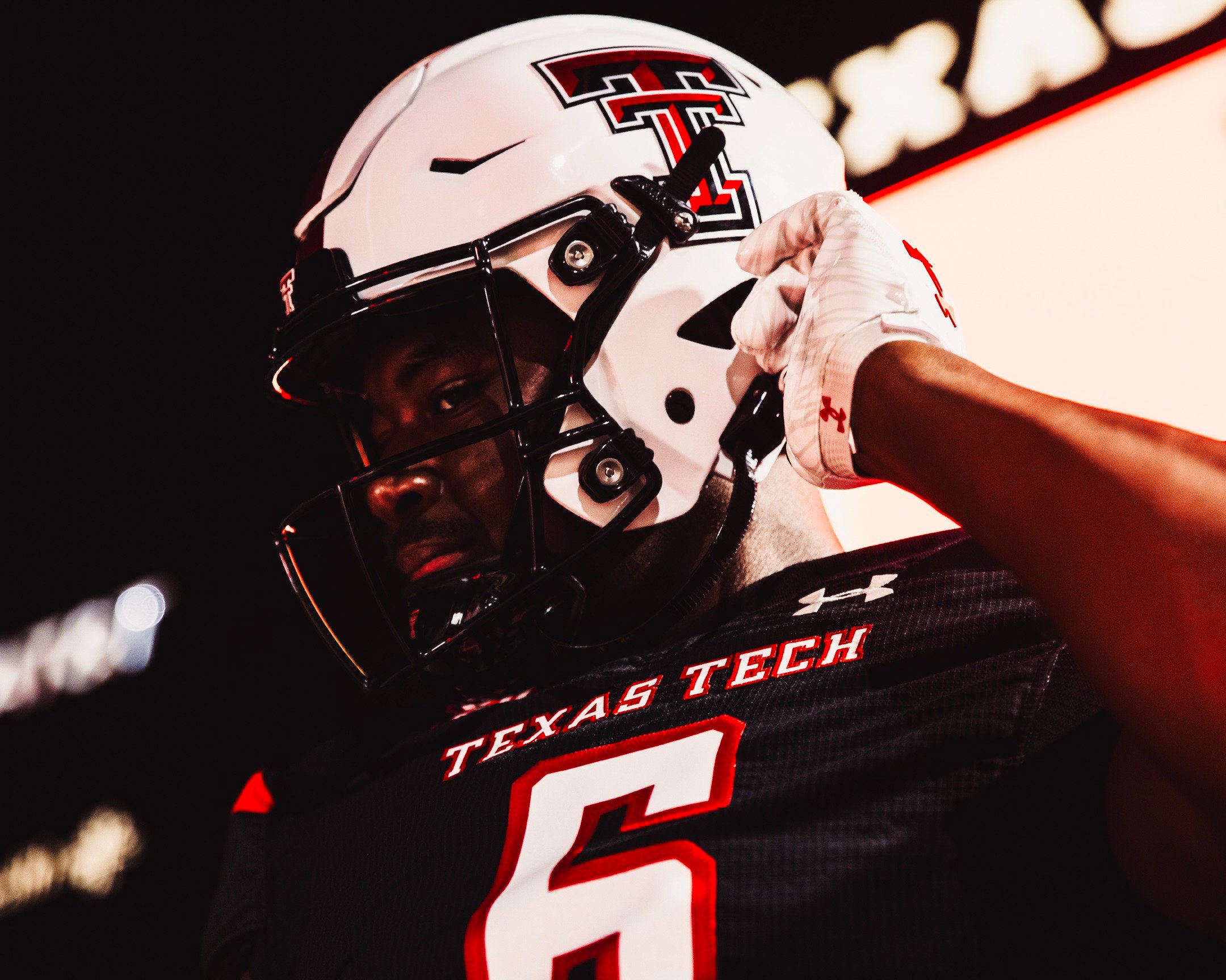 A football player from Texas Tech wearing a black jersey with the number 5 and a white helmet with a Texas Tech logo is adjusting his helmet.