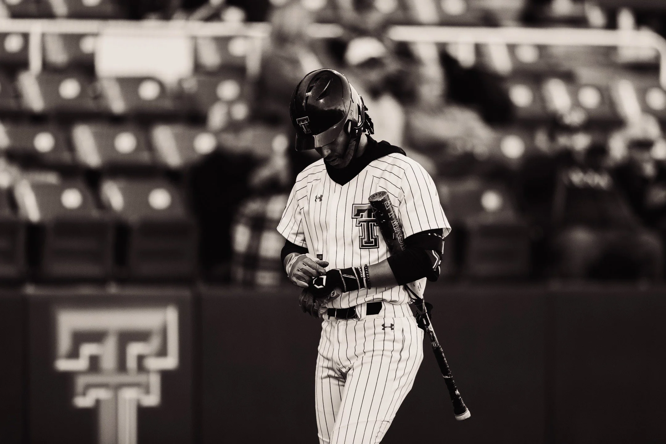 A baseball player in a uniform with pinstripes standing on the field with a bat hanging from his back, looking down at his wrist, in a stadium with empty seats in the background.