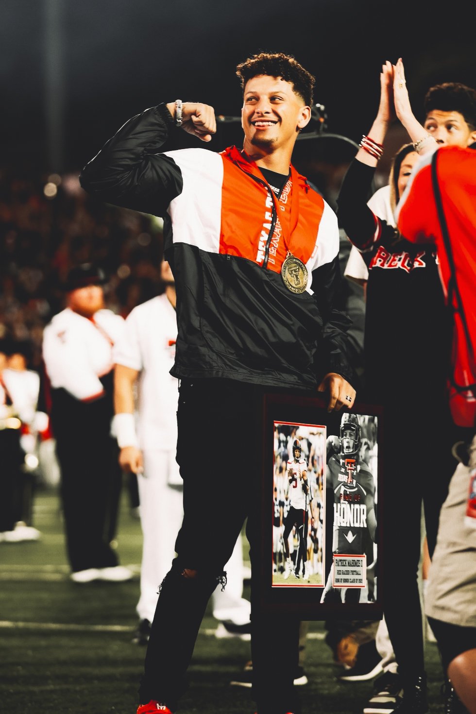 Young male athlete at an award ceremony on a football field, wearing a jacket with a gold medal around his neck, flexing his arm, and holding a framed photograph of a football player.
