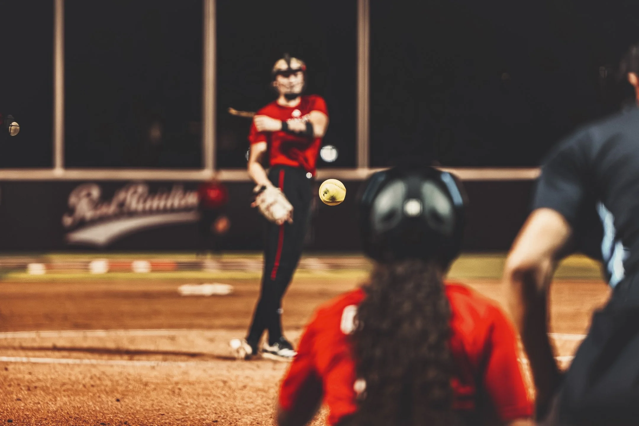 Female softball player in red and black uniform throwing a yellow softball during a game at night, with a catcher in red gear in the foreground and another player in black on the right.