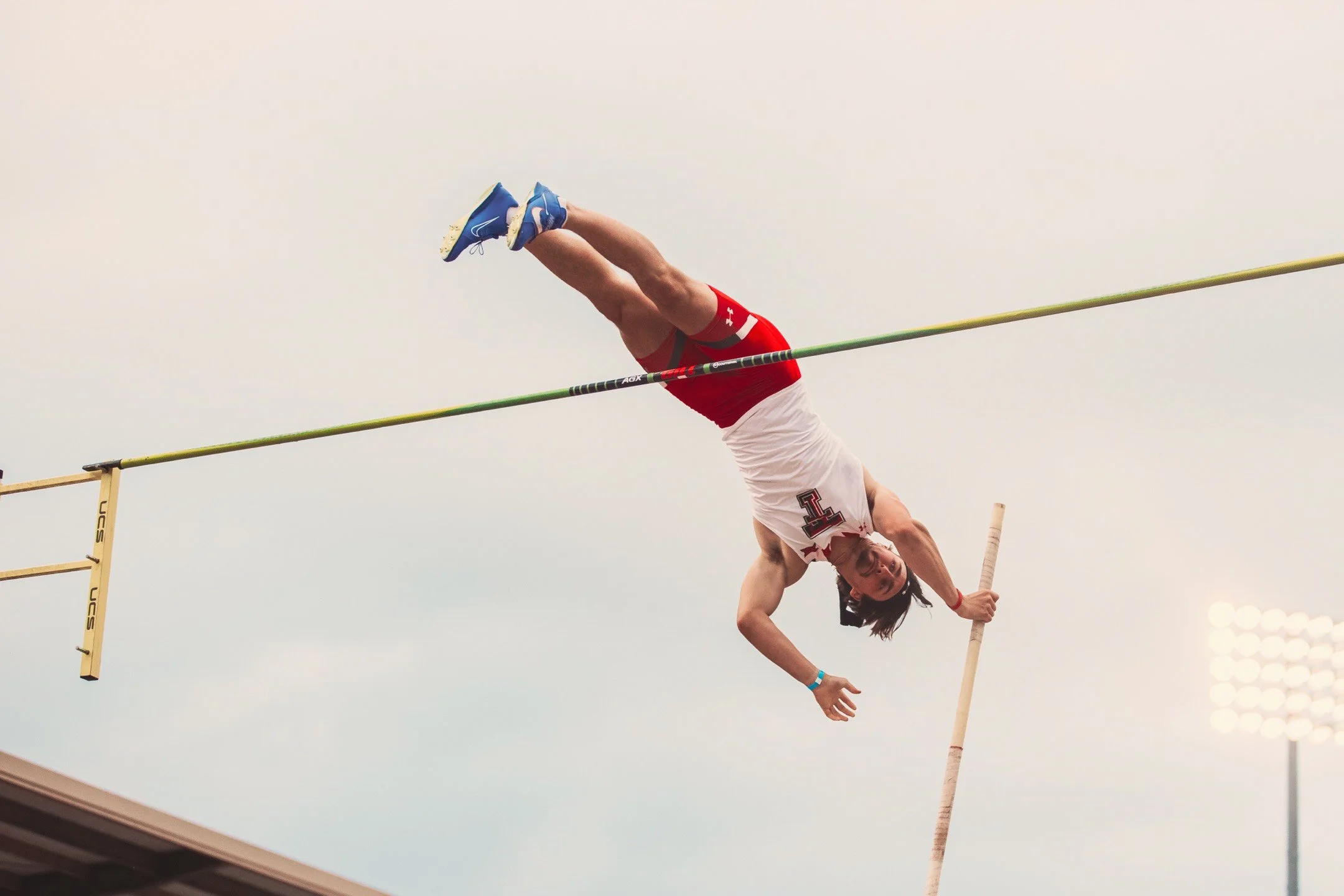 Male pole vaulter in red and white uniform mid-air during vault attempt at an outdoor track and field stadium.