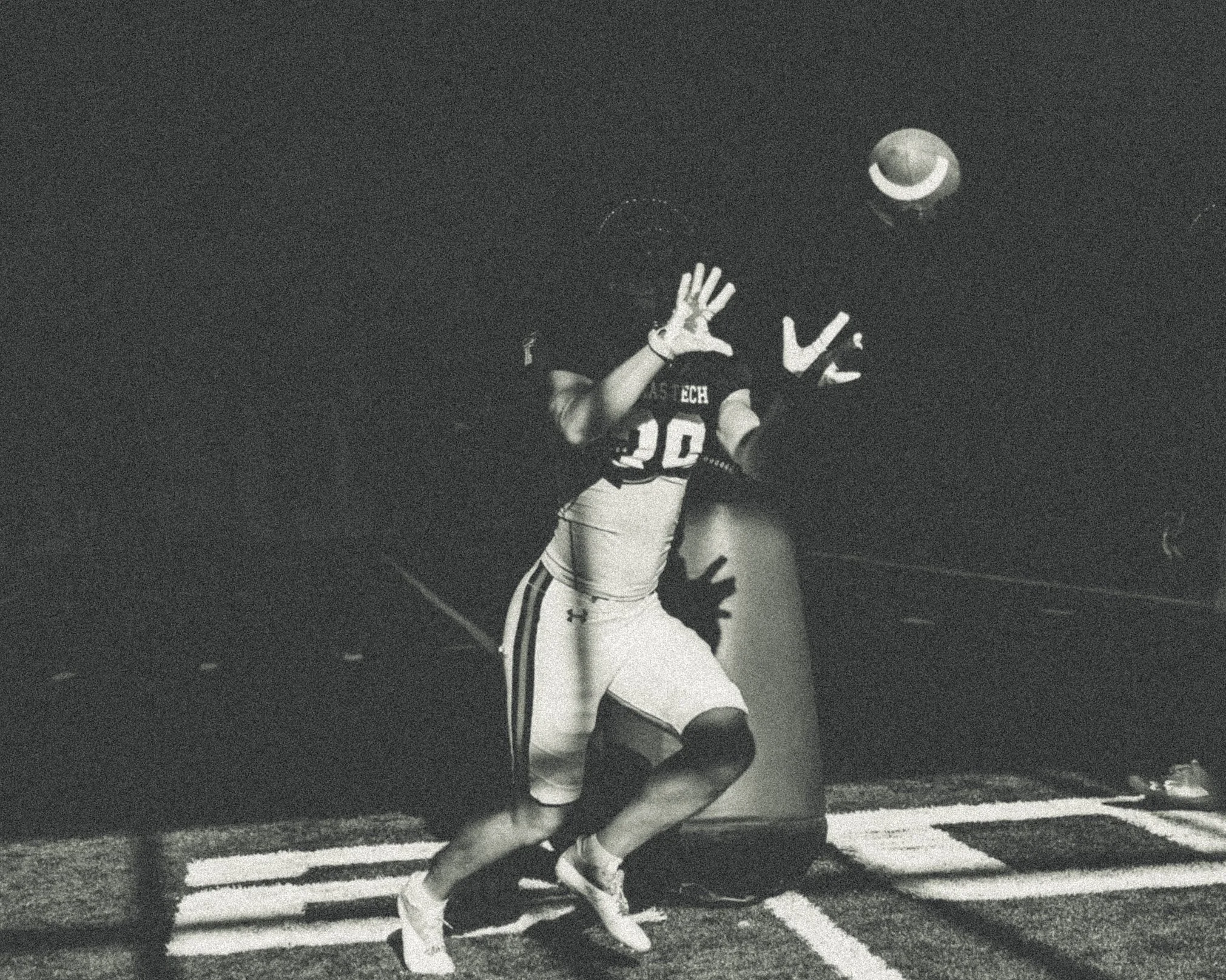 A football player attempting to catch a football in a dark stadium, with his hands raised and eyes on the ball.