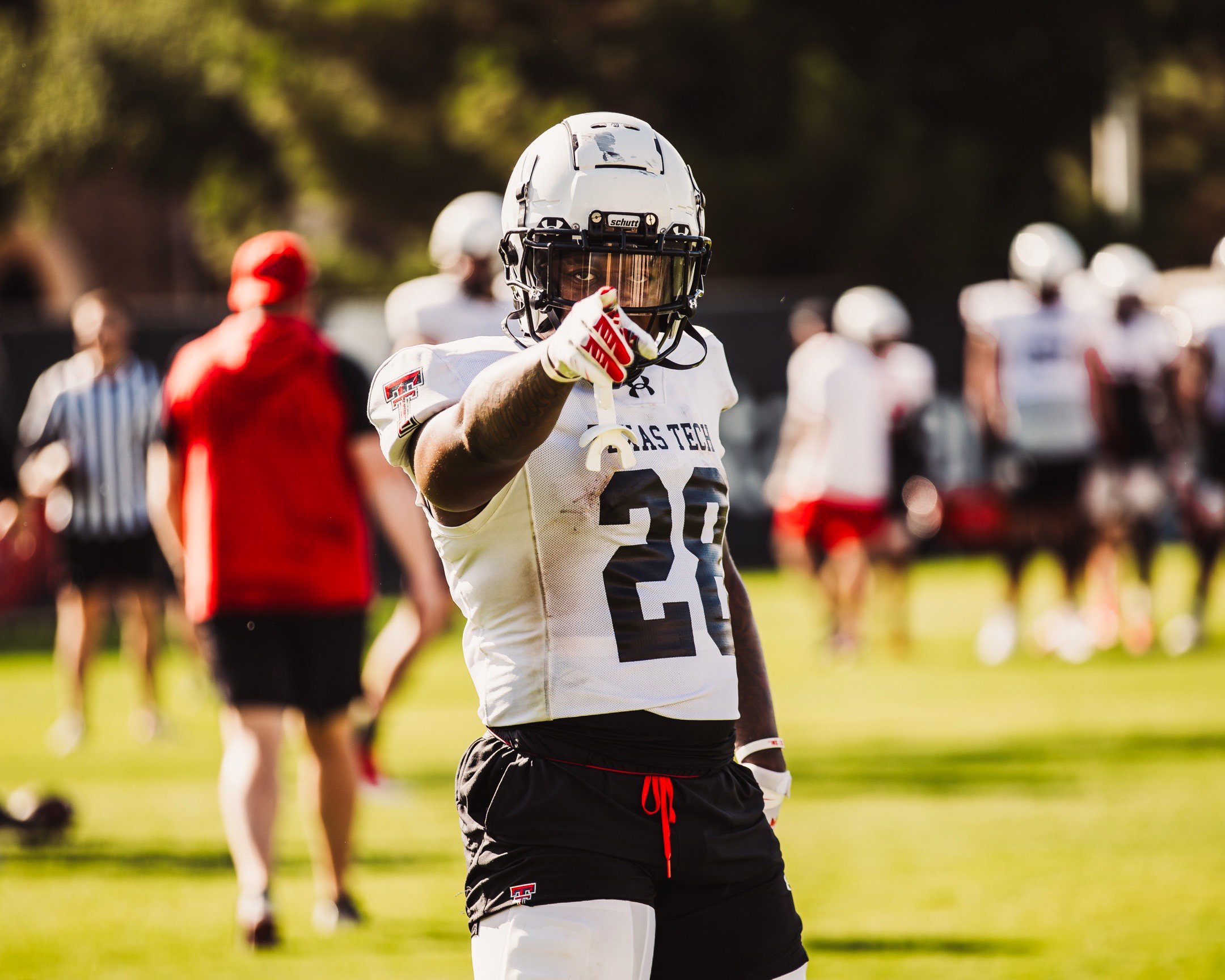 An American football player in a white jersey and black shorts on the field pointing at the camera during a game or practice.