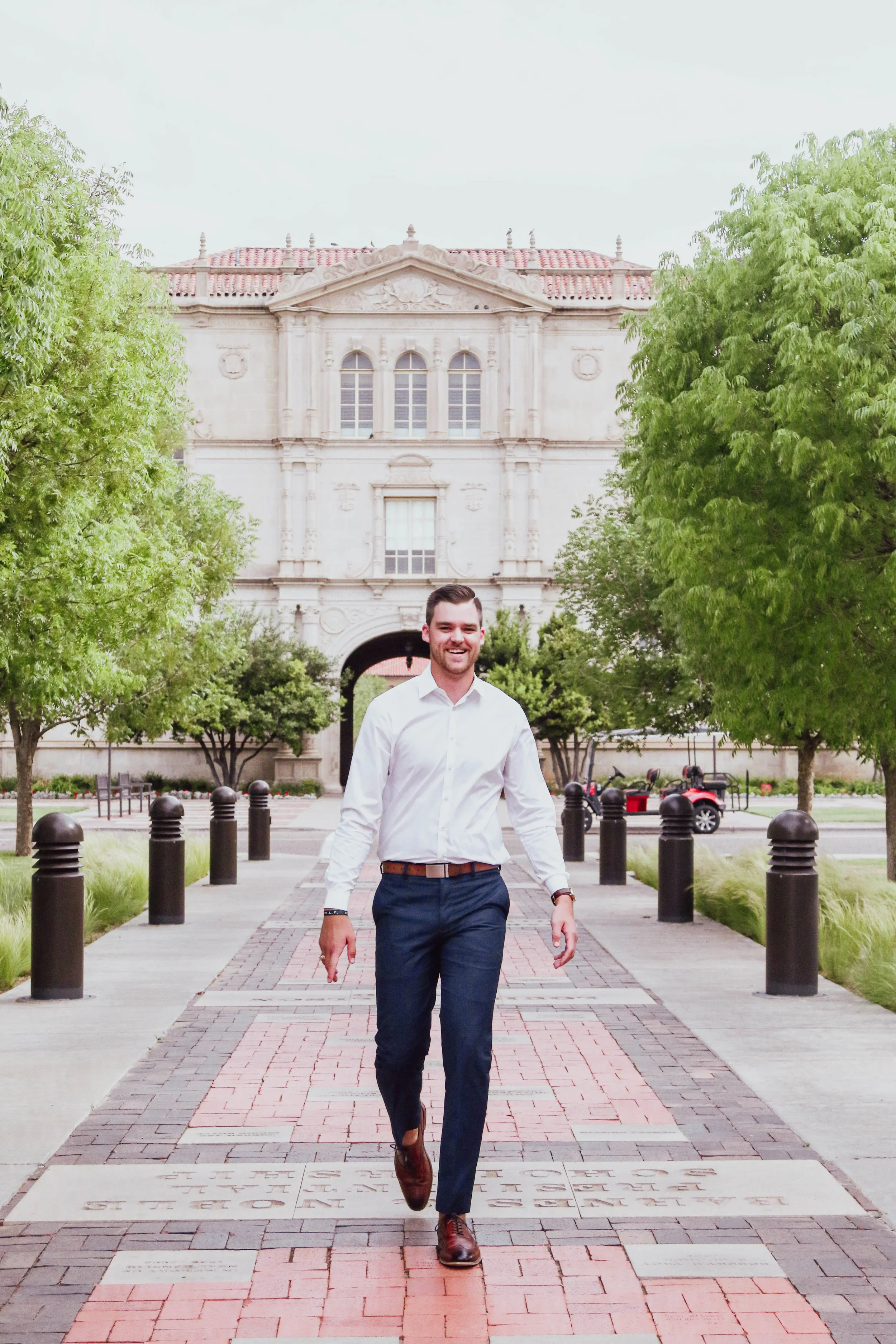 A man in business attire walking on a pathway in front of a historic building with trees flanking the path.