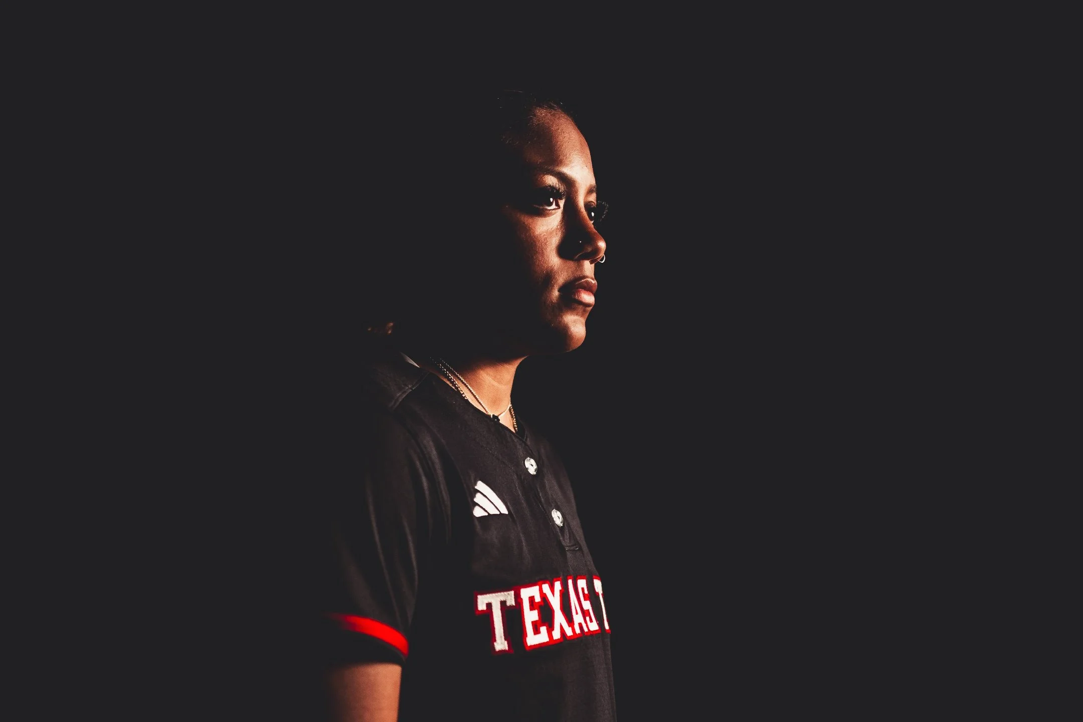 A woman with dark skin, short hair, and hoop earrings, wearing a black sports jersey with 'TEXAS' written on it, looks to her left against a dark background.