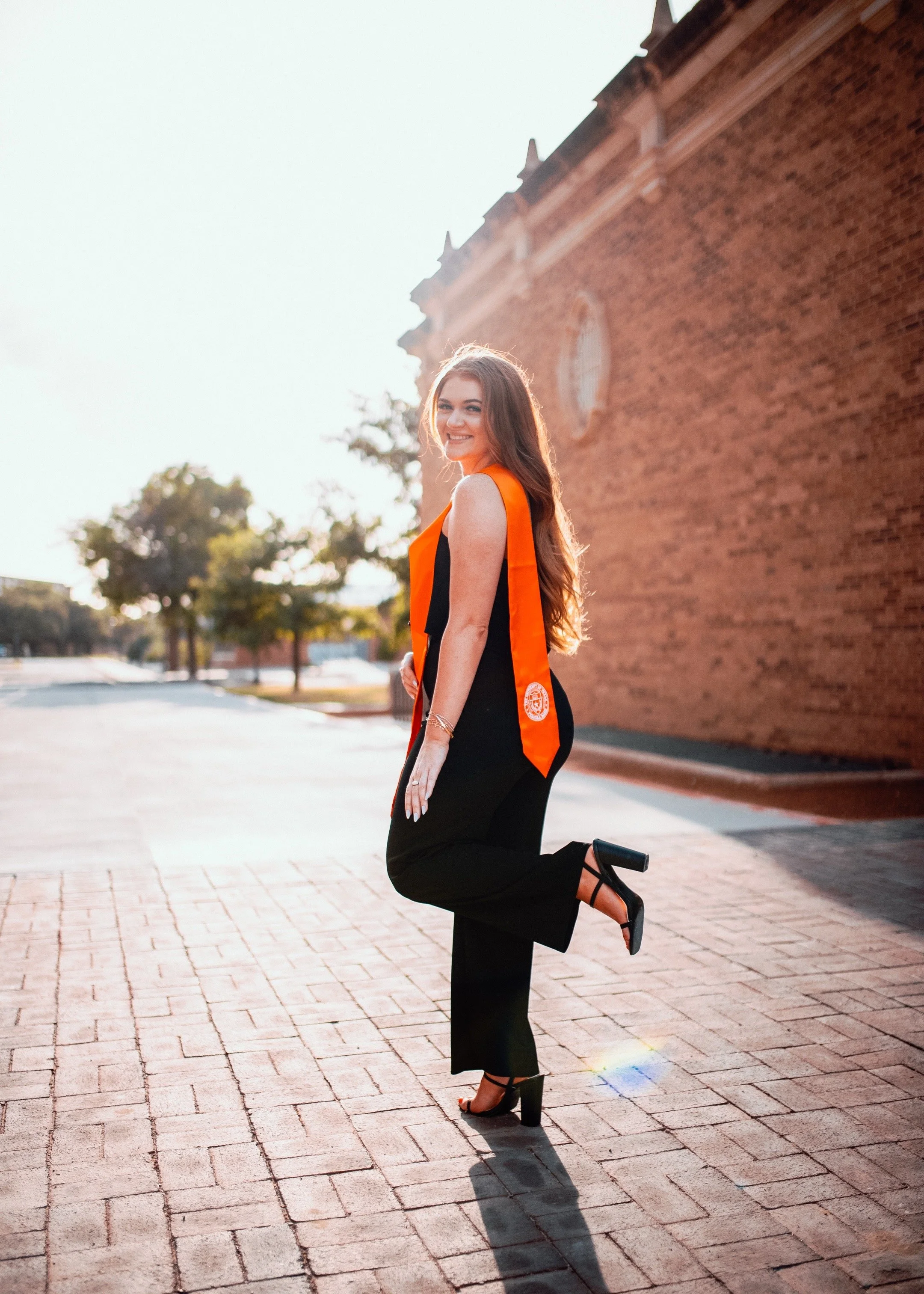 A young woman in black dress and high heels standing outdoors, smiling, with a brick building in the background, wearing an orange graduation stole.