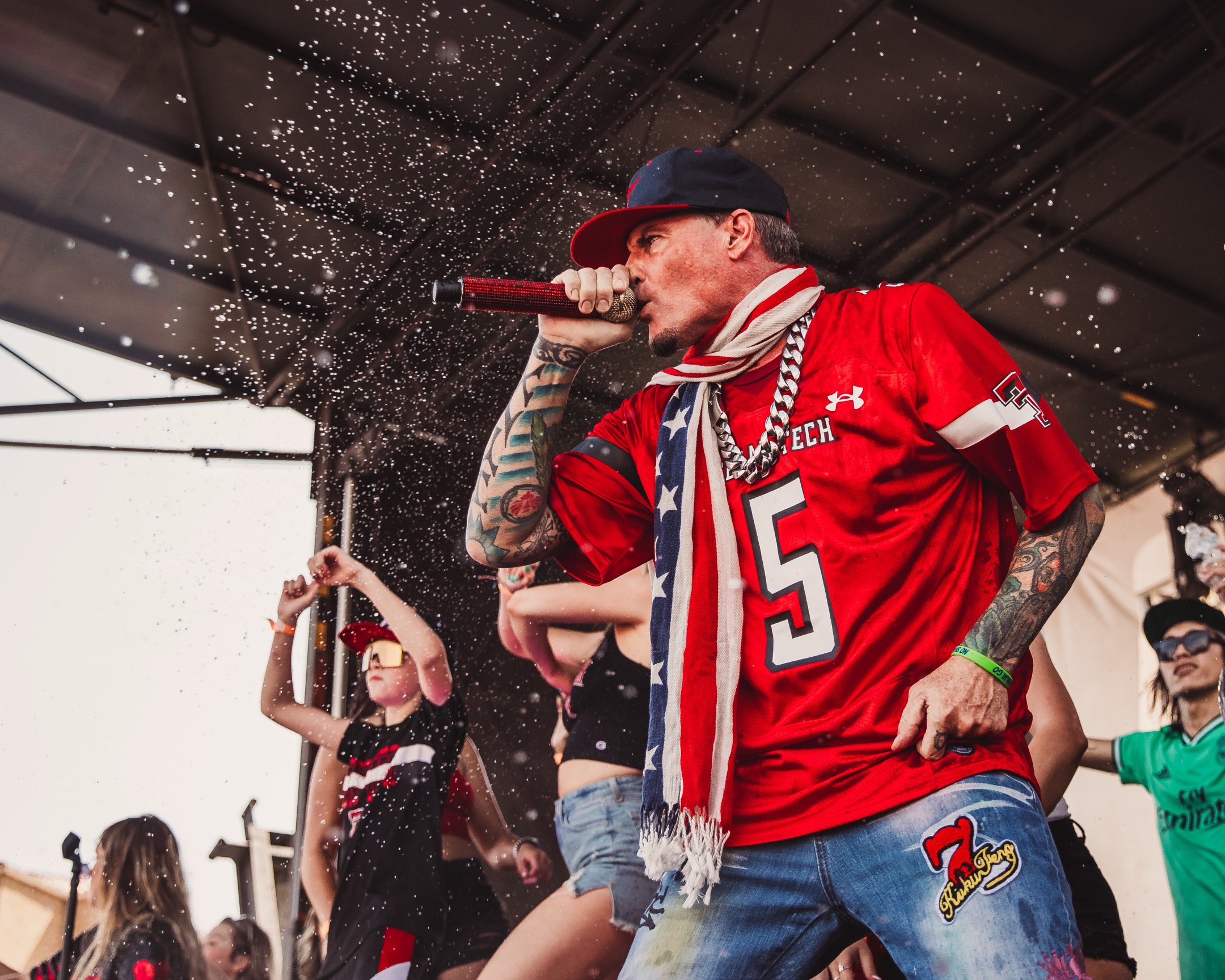 A man with tattoos singing into a microphone on stage, wearing a red sports jersey and a scarf around his neck, with people dancing in the background.