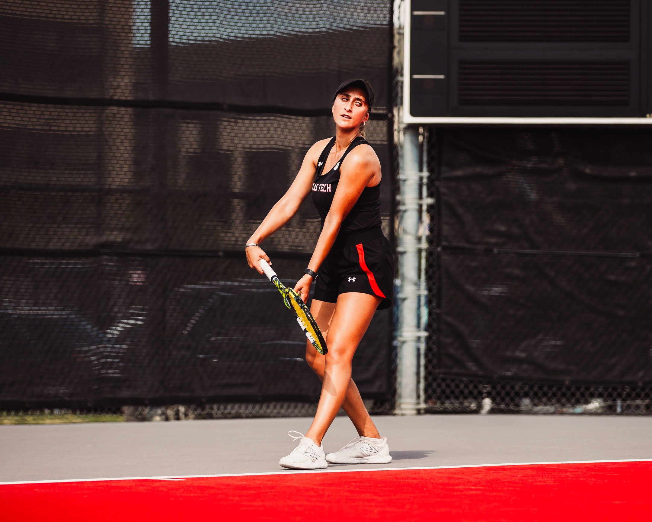 A woman in athletic wear holding a tennis racket on a tennis court with black wind screens in the background.