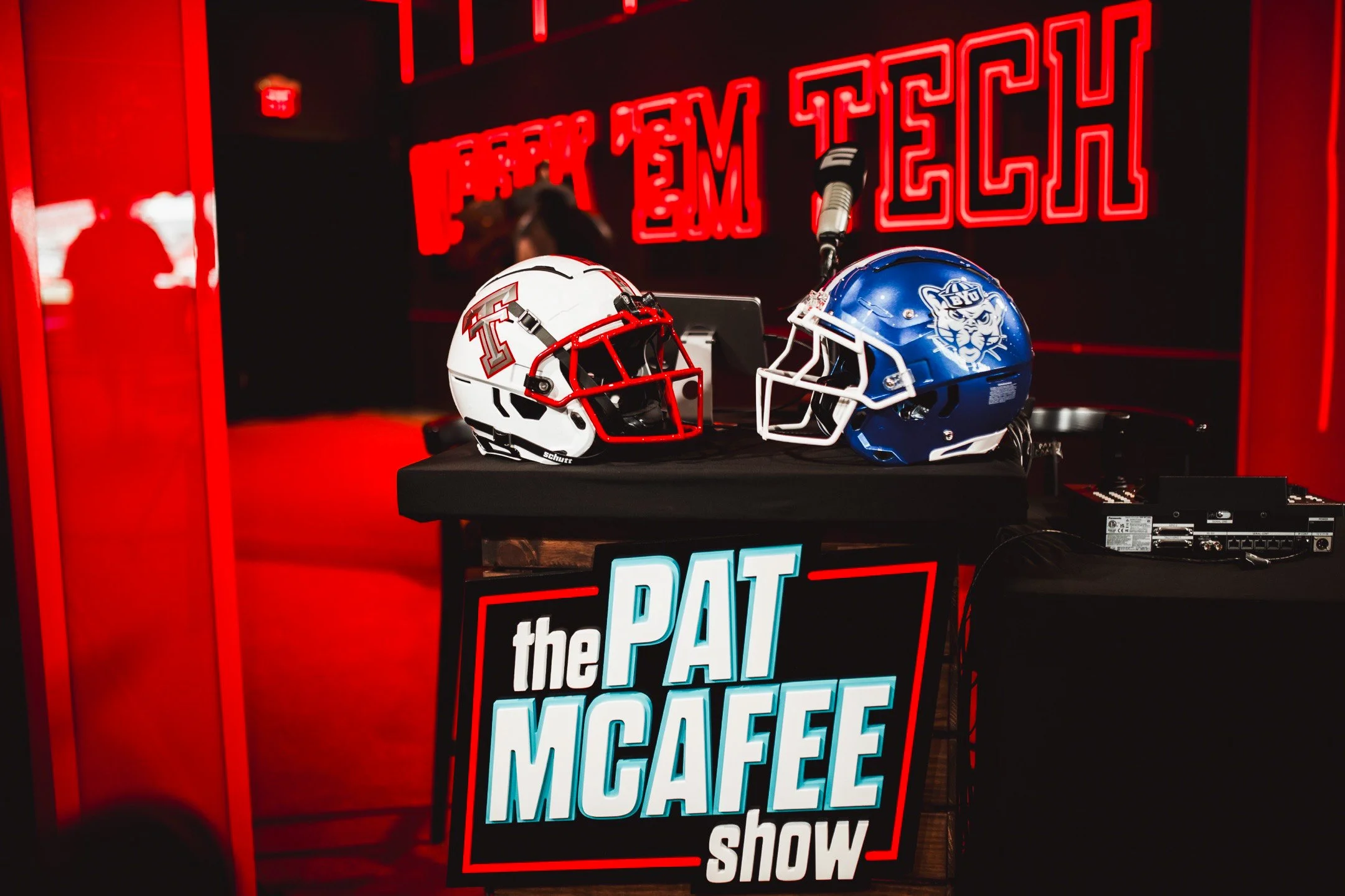 Two football helmets, one with Texas Tech logo and the other with a blue panther face, are displayed on a table with a sign reading 'the PAT McAFEE show'. In the background, there is a red neon sign that says 'FROM THE TECH'.