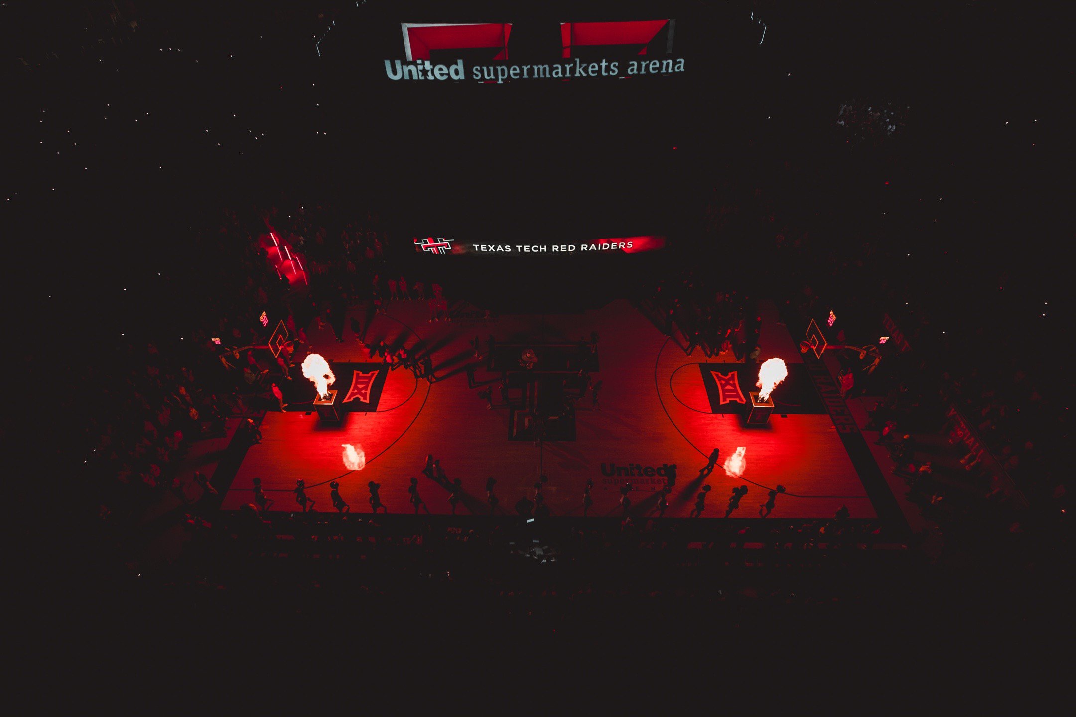 An indoor basketball court with a red-lit floor, featuring the Texas Tech Red Raiders logo at the center. Flammable pyrotechnics are on the court, with flames shooting up near the team's logo. Above the court, a digital screen displays the text 'Unit