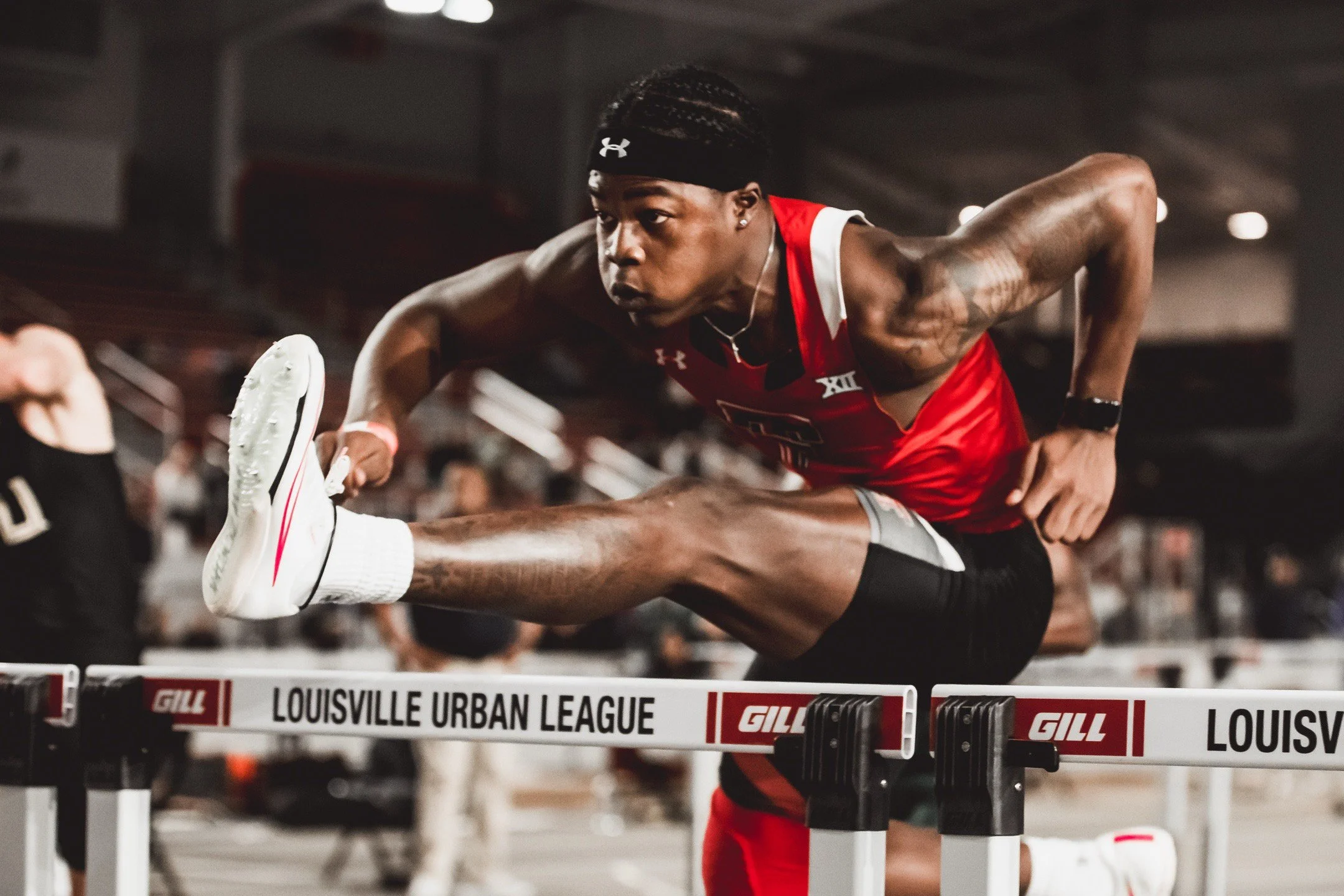 An athlete wearing a red singlet and black shorts is competing in a hurdle race, leaping over a hurdle labeled 'Louisville Urban League' during a track and field event.