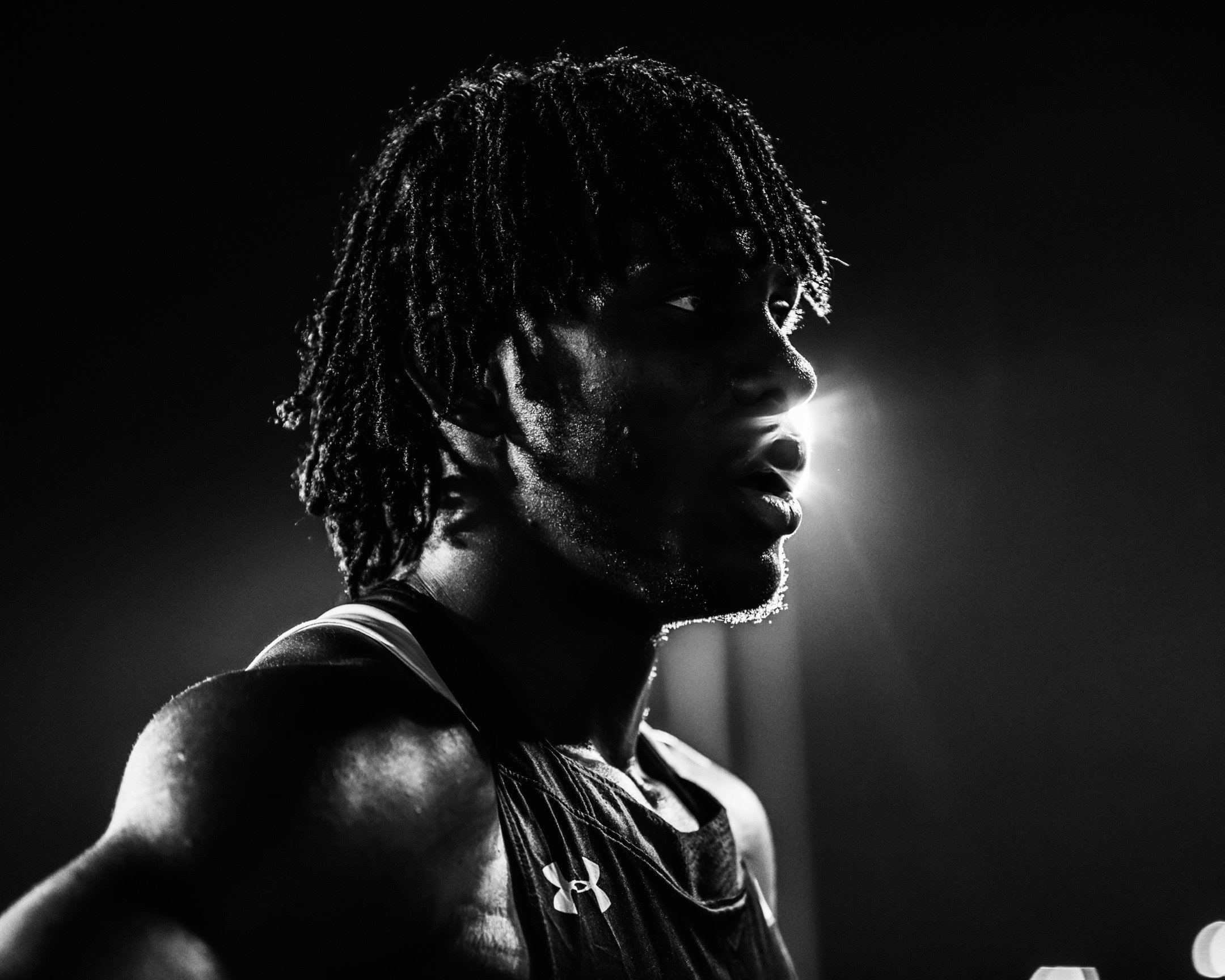 A black-and-white photo of a young male athlete with dreadlocks, sweating, looking to the right, illuminated from behind with a bright light source.
