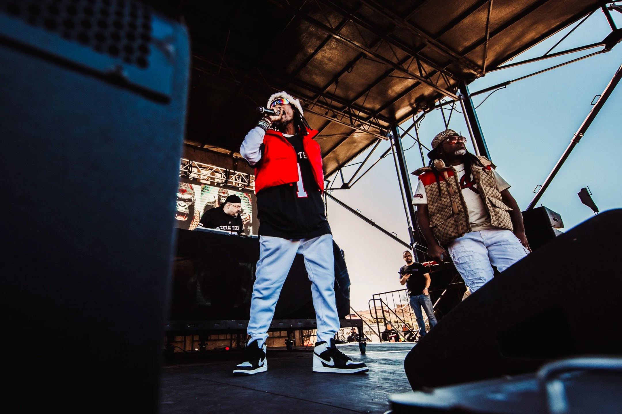 Musicians performing on an outdoor stage, with a DJ and a man in sunglasses in the background, under a large metal canopy during sunset
