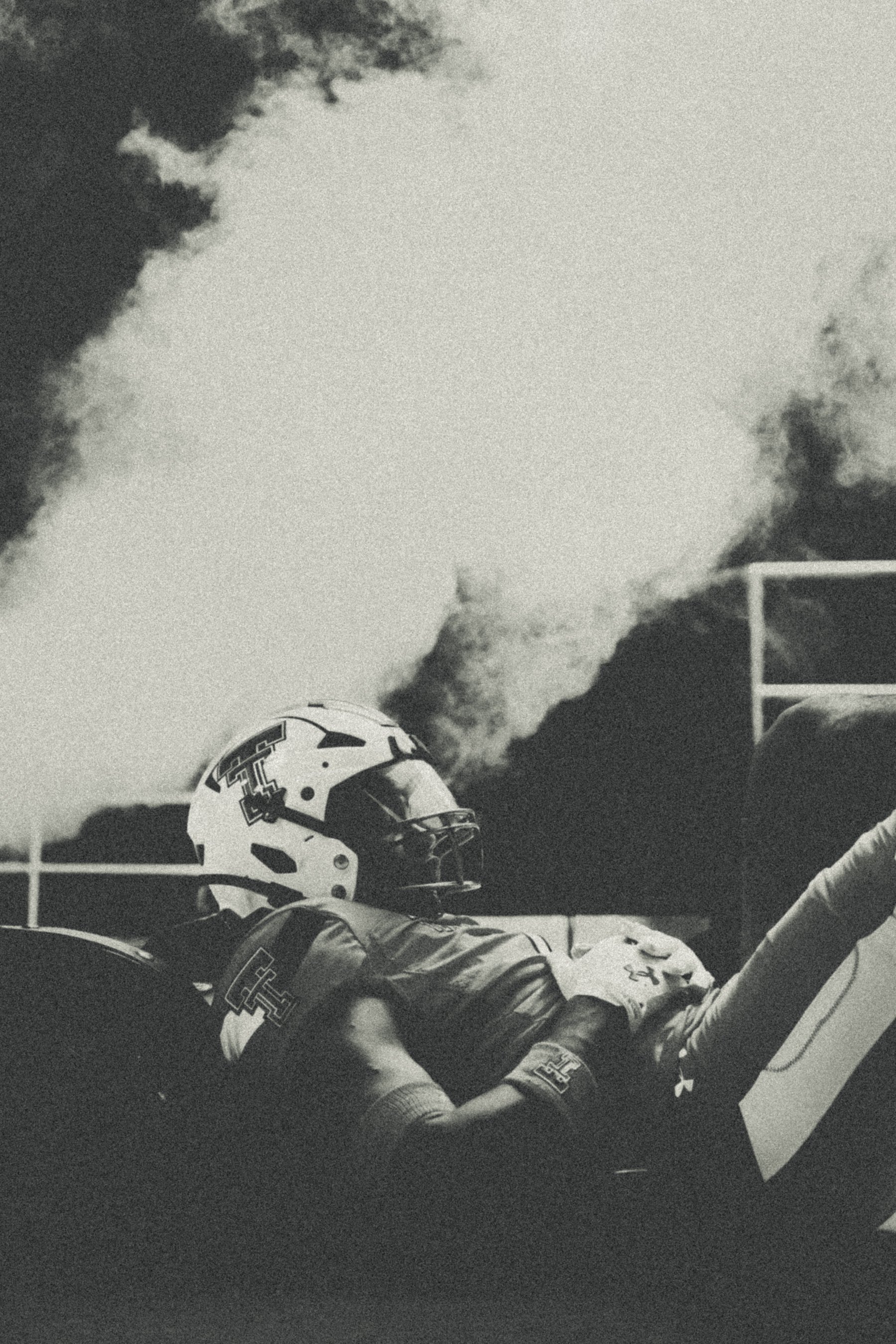 A person in a racing helmet and suit sitting in a race car on a track, with smoke or clouds in the background.