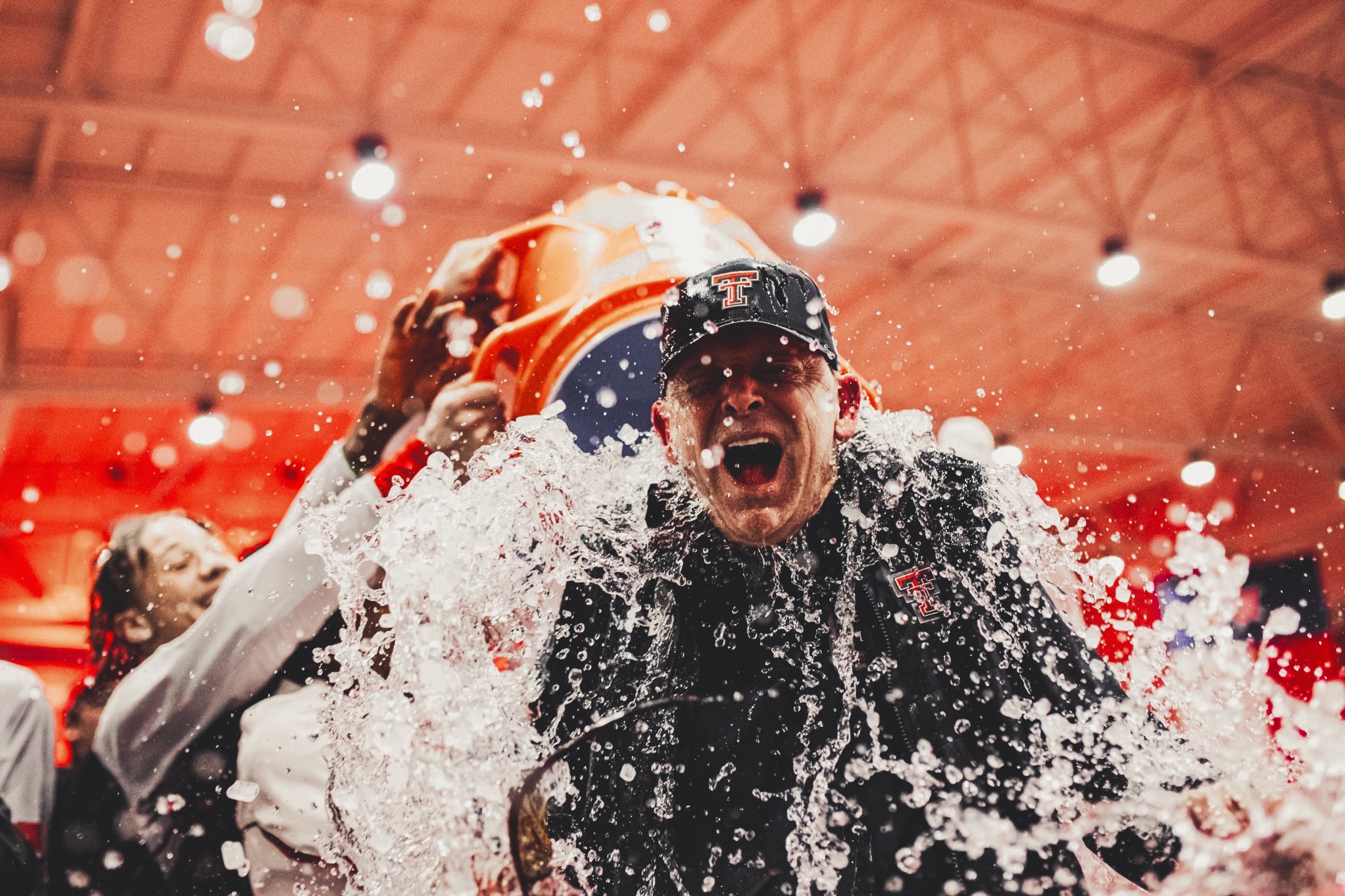 A man wearing a black Texas Tech hat and jacket, celebrating with water splashing around him, holding a helmet above his head. A woman in the background is also celebrating.