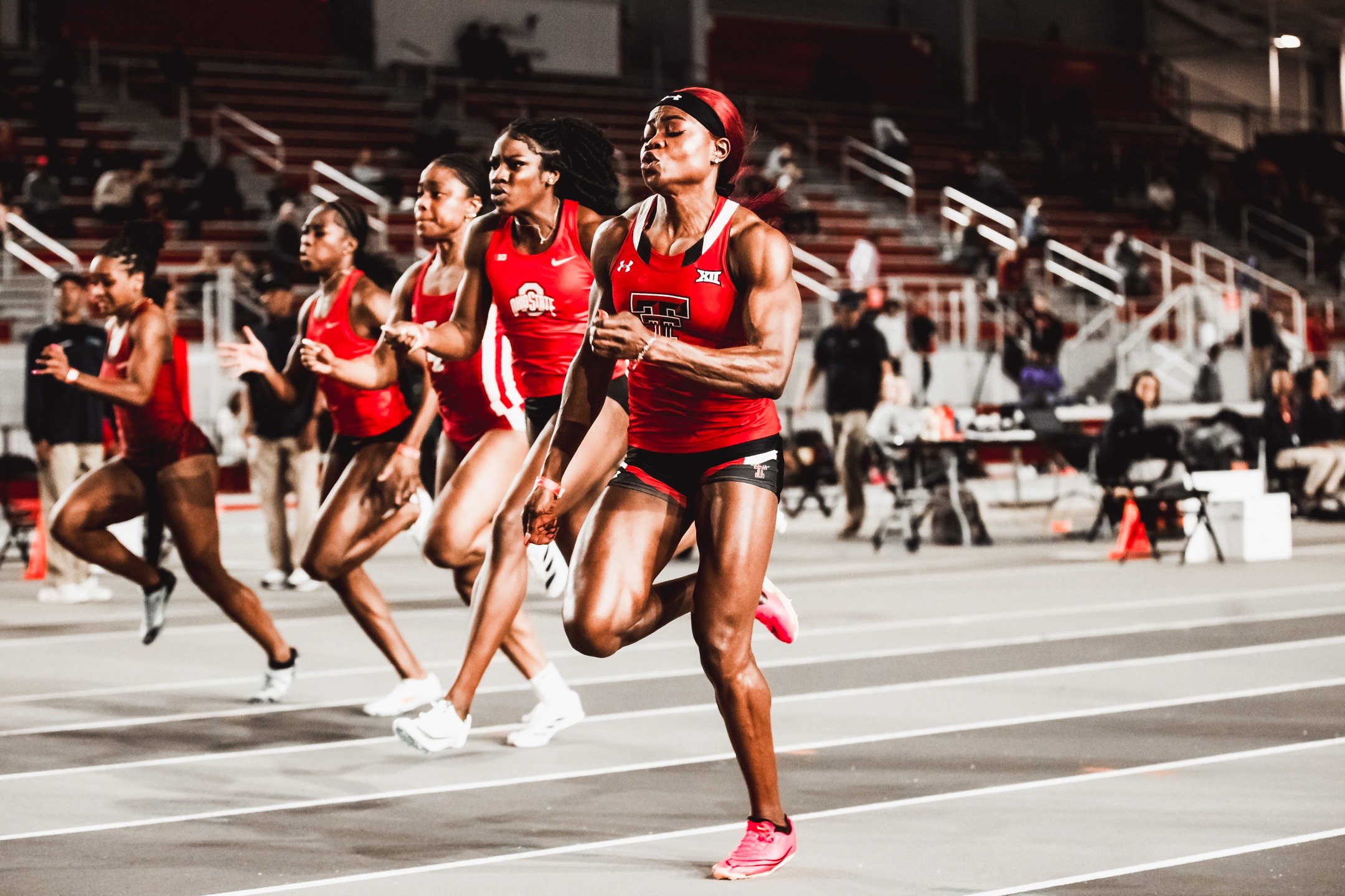 Female athletes in red uniform sprinting on a track during a race at a stadium.