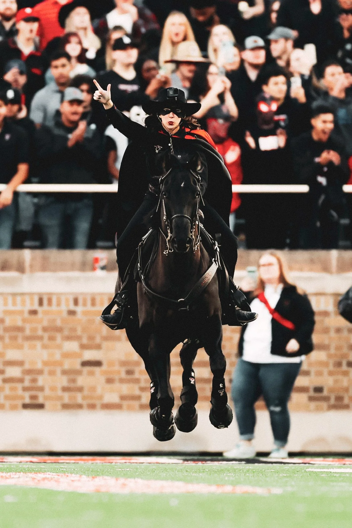 A woman in black riding attire, wearing a hat and cape, rides a black horse and raises her right hand with two fingers extended during a football game at Jones AT&T Stadium in Lubbock, Texas.