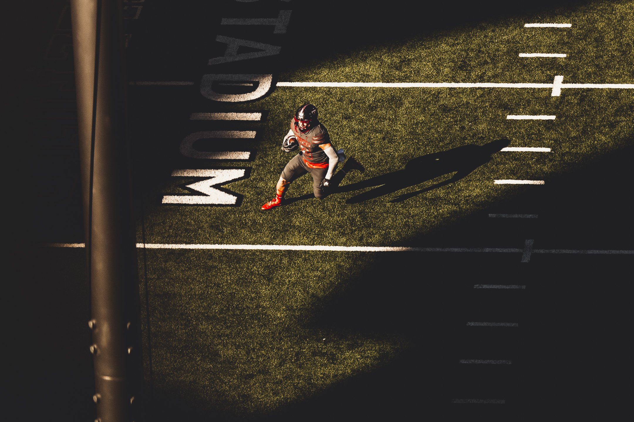 Top-down view of an American football player in a black and orange uniform running on a green turf field with white yard lines, casting a long shadow.