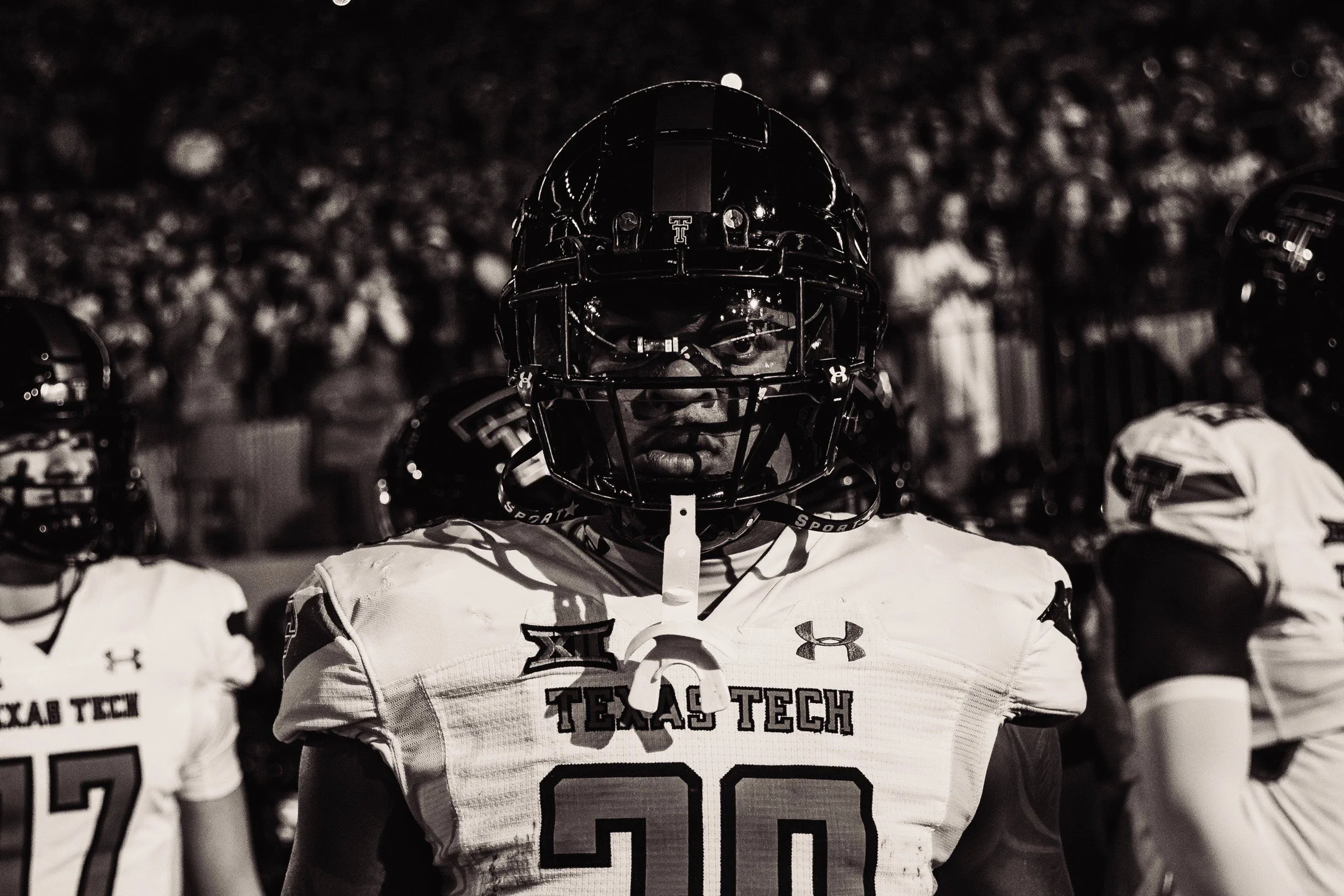 A football player wearing a helmet and jersey with the number 20, standing on the field during a game with team Texas Tech, with other players and a crowd in the background.