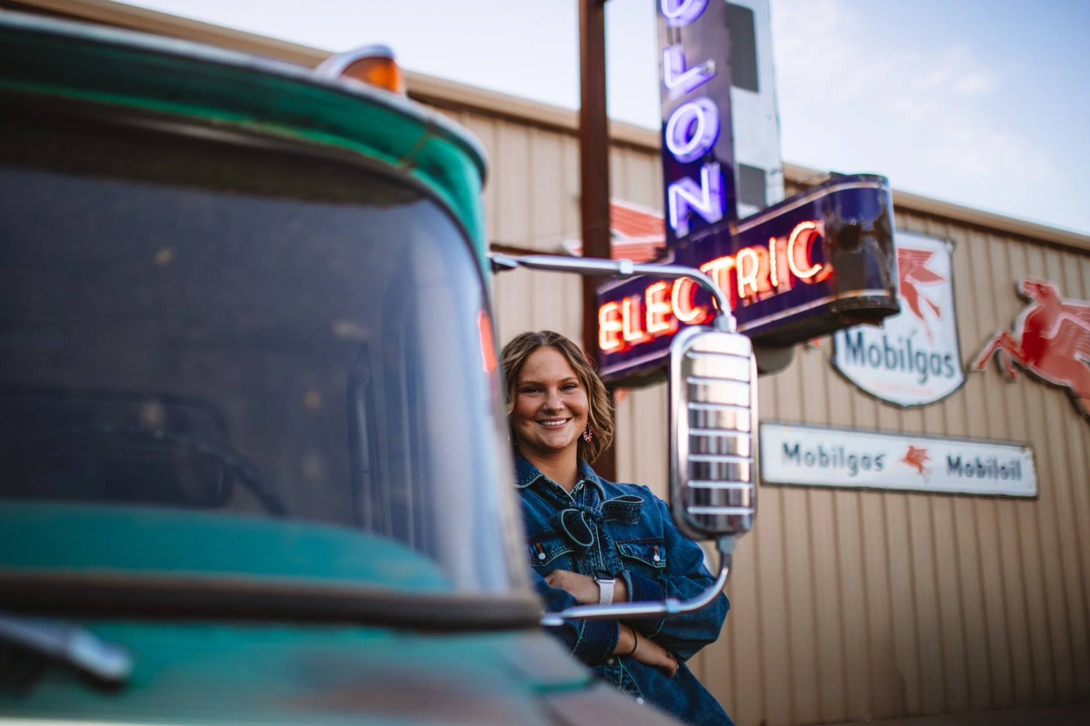 A woman smiling and leaning against a turquoise vintage vehicle in front of an electric sign and a Mobilgas sign outside a building.